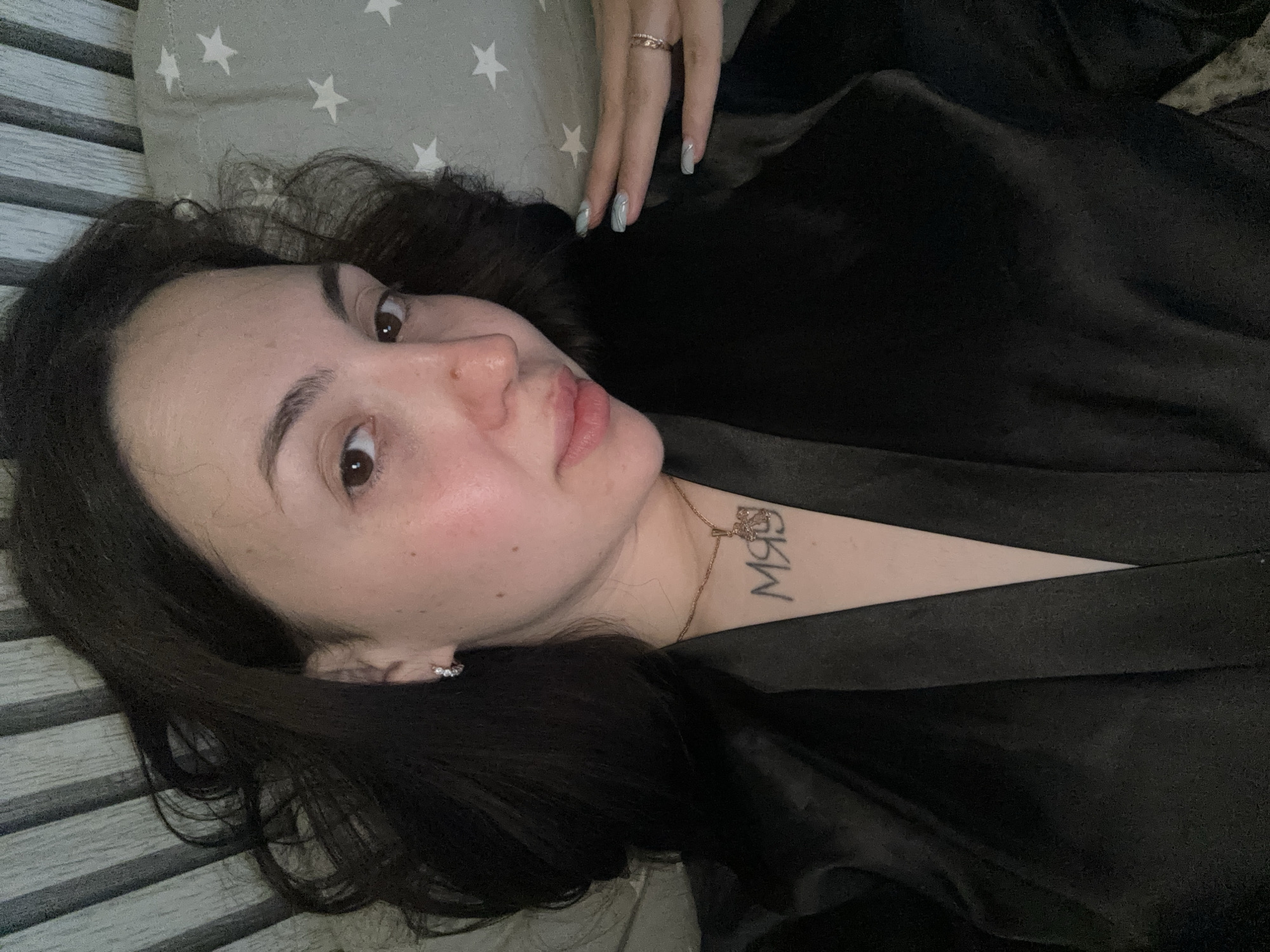 A close-up, slightly overhead view depicts a woman lying down indoors in Марковский, Russia. She has dark, wavy, shoulder-length hair and fair skin with visible natural texture and some light redness on her cheeks. Her eyes are dark brown, and she wears a subtle, relaxed expression with a direct gaze towards the camera.
She is adorned with small, clear-stoned hoop earrings in her left earlobe, a thin gold-colored chain necklace with an intricate pendant, and a stone-set band ring on the ring finger of her right hand. Her nails are manicured in a light blue/mint green shade with white swirl designs on several digits. Prominently visible on her sternum, below the collarbone, is a tattoo of Cyrillic script reading "МЯУ" (MYAU). She is dressed in a dark, possibly black or charcoal, V-neck garment, likely a satin or silk robe, which reflects light slightly.
The background to her left consists of vertical striped paneling in shades of grey and off-white. To her right, a light grey fabric with white star patterns is visible, suggesting a pillowcase or bedding. Her right hand is gently raised, with fingers resting on this starry fabric near her neck.