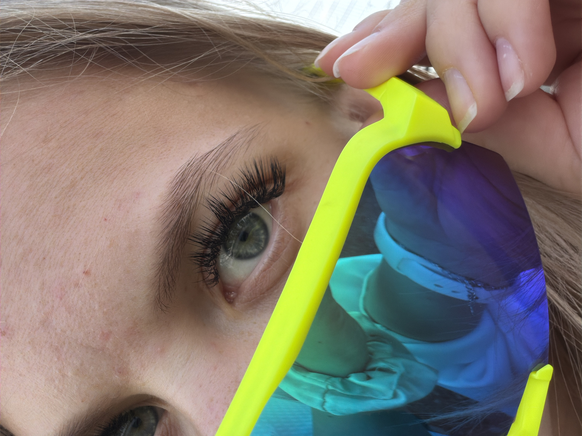 A close-up shot focuses on a person's eye, peeking out from behind bright yellow sunglasses with iridescent lenses that reflect a blue and green gradient. The person's eye is a striking blue-green, with long, dark eyelashes framing it. The skin around the eye shows subtle freckles and a few small blemishes. A hand with neatly trimmed nails is visible on the right side of the frame, holding the sunglasses. Light-colored hair is visible on the right side of the image. The background is out of focus but suggests an outdoor setting. The image appears to be taken in Chaikovsky, Russia, as indicated by the provided information.