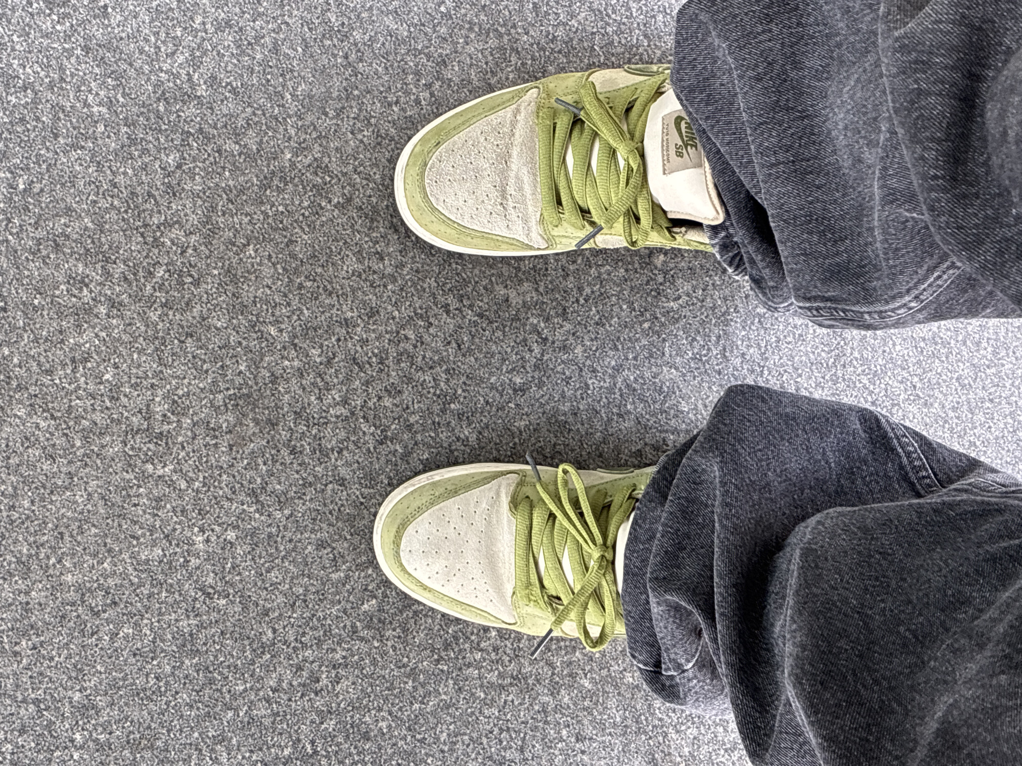 From an elevated perspective, the image features a person's lower legs and feet, positioned on a gray, grainy surface resembling concrete or asphalt. The individual is wearing faded black or dark gray denim jeans, which are slightly wrinkled around the knees. The footwear consists of two Nike SB sneakers, predominantly in a pale green hue, with white accents on the toe box and the tongue label. The laces are also green and intricately tied. The composition is framed to exclude the upper body and broader surroundings, focusing primarily on the lower portion of the person and the ground. There is no indication of Barcelona, Spain in the image.