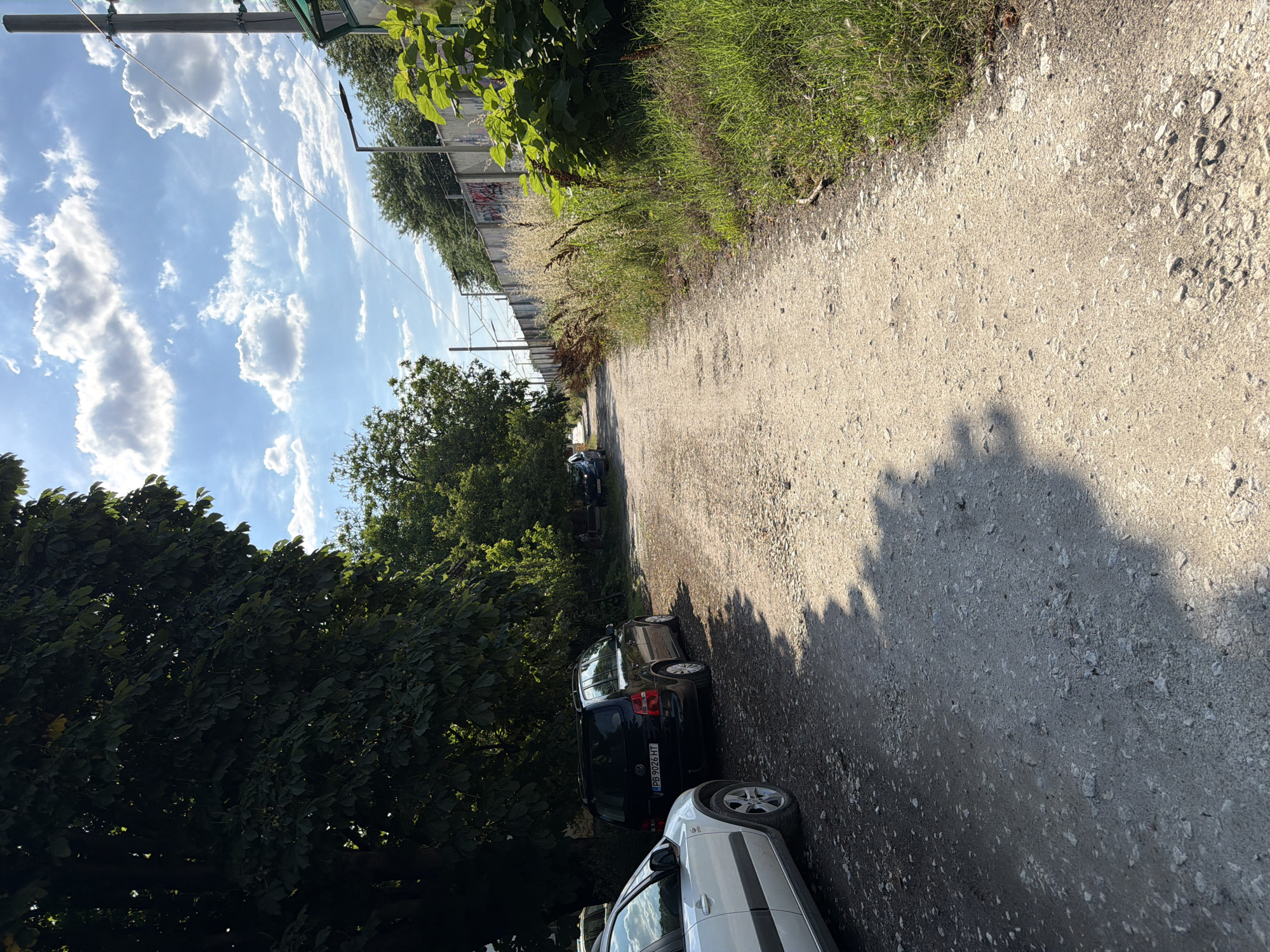The image depicts a gravel road lined with trees on the left and tall grasses and a concrete barrier with graffiti on the right. Several parked cars are visible on the left side of the road, including a white vehicle in the foreground and a black Volkswagen Touran. The road appears to be unpaved and slightly dusty. In the background, power lines and poles stretch along the right side of the scene, suggesting the proximity of infrastructure. The sky is blue with scattered white clouds, indicating fair weather. In the distance, on the right, a small group of people can be seen sitting near some trees. The scene is likely located in an area bordering a more developed part of the city, possibly near a transportation route or industrial zone. The overall impression is one of a quiet, somewhat undeveloped, outdoor space within an urban environment.