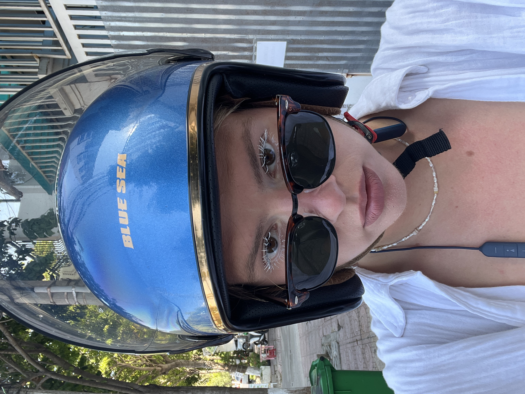 A person wearing a blue motorcycle helmet labeled "BLUE SEA" and sunglasses is positioned in Nam Nha Trang Ward, Vietnam. The individual is wearing a white shirt and a beaded necklace with a dark corded electronic device. The surrounding environment includes trees, a green trash receptacle, and a building with a corrugated metal awning.
