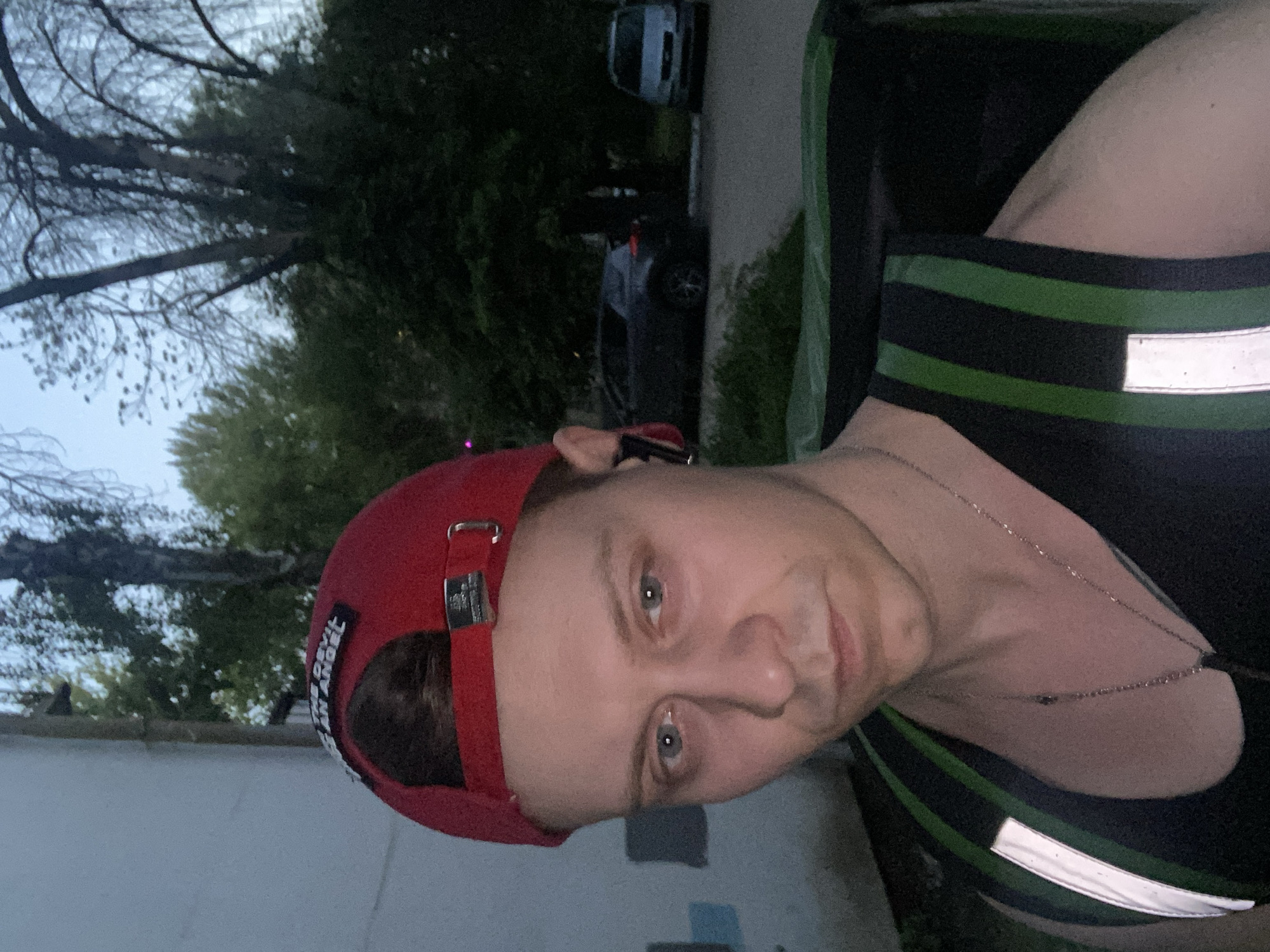 A man is standing in front of a building, possibly at dusk or dawn. He is wearing a red baseball cap with text that reads "DEVIL AND ANGEL". He has on a black tank top with a green and black striped vest over it, which has reflective strips. He is wearing a silver chain necklace. In the background, there are trees and parked cars, one of which is a light-colored sedan and another a dark-colored SUV. The scene appears to be an urban setting in Samara, Russia.