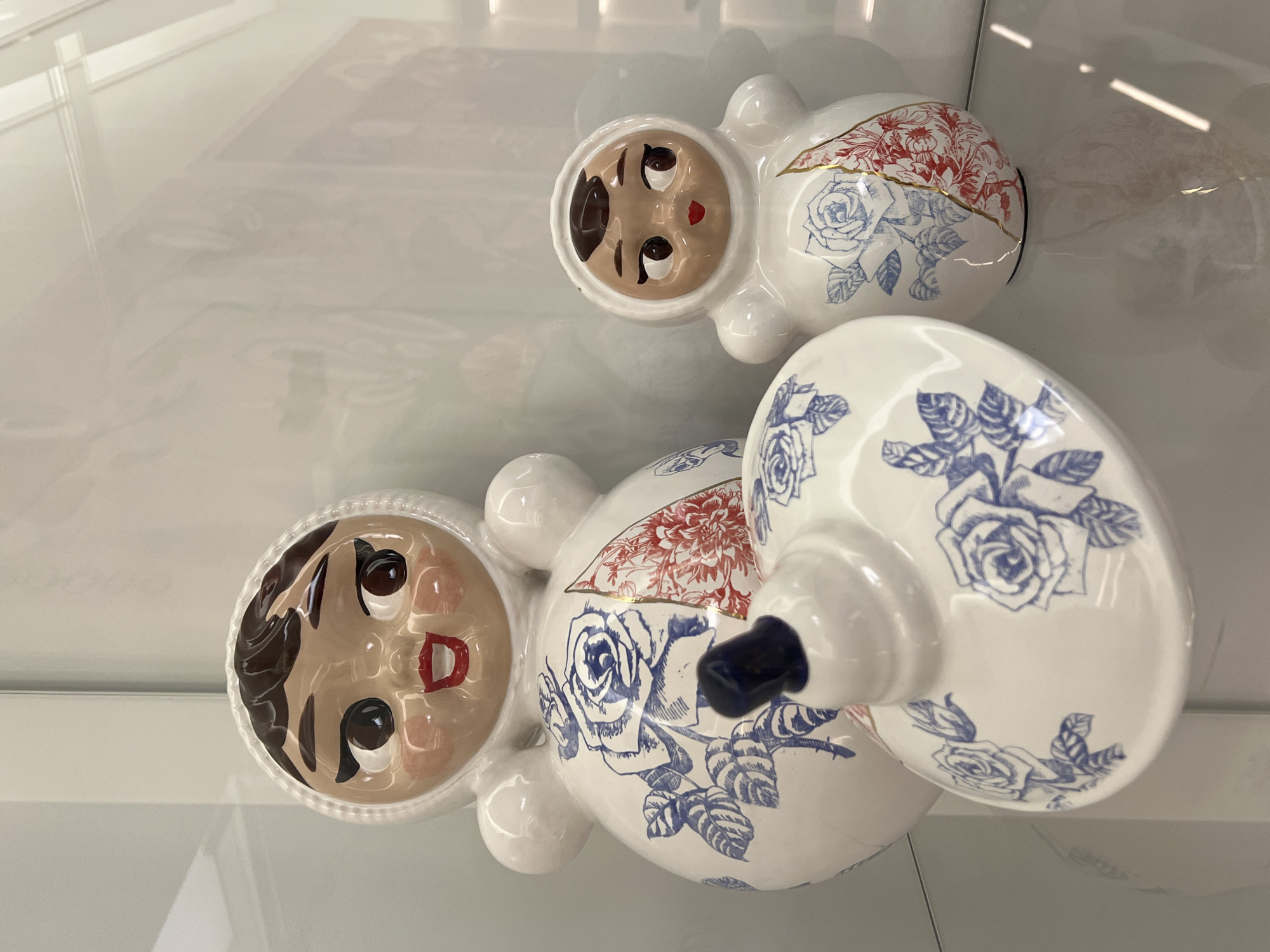 The image shows two ceramic figures, seemingly displayed within a clear, reflective case or shelf, potentially made of glass or acrylic. The figures are doll-like, with painted faces and rounded bodies. They are primarily white, decorated with blue and red floral patterns. The larger figure has a tiered design, with a broader base that tapers into a smaller upper section. The location is in the city of Unknown, Unknown.