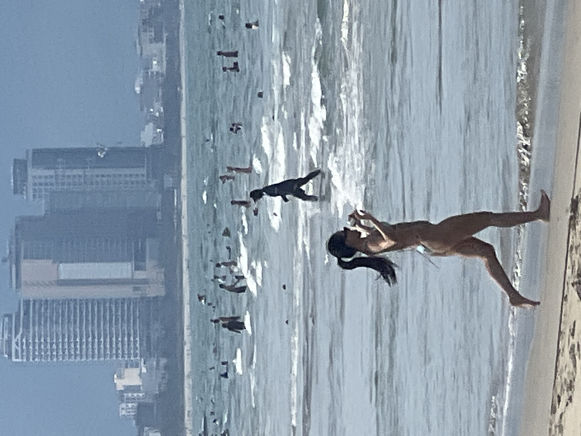 The image captures a sunlit day at a beach in Da Nang, Vietnam. The scene features several people in the ocean and on the shore. In the foreground, a woman in a bikini is standing in the shallow water, looking down at her phone. Her dark hair is tied in a ponytail. Further out in the ocean, a dark-colored dog is swimming. Numerous other figures, likely people, are scattered throughout the water, some appearing to be playing or wading.
In the background, a hazy cityscape with tall buildings is visible, suggesting an urban coastal environment. The ocean is calm with gentle waves. The lighting indicates it is daytime, and the bright atmosphere suggests clear weather. The overall mood appears relaxed and leisurely, typical of a beach day. No visible text is present in the image.
