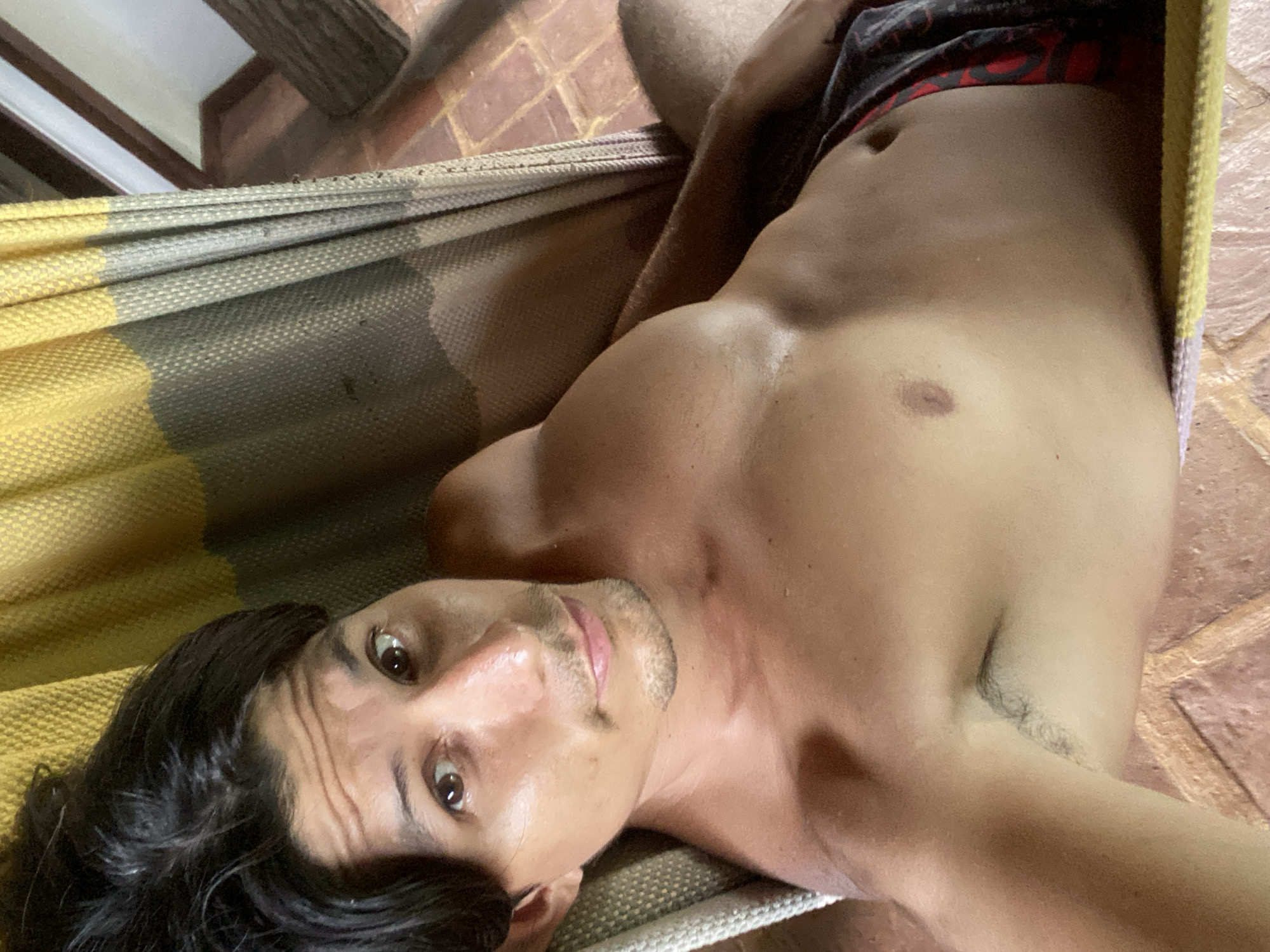 The image shows a person lying in a hammock. The person is shirtless, wearing patterned shorts, and has dark hair and brown eyes. Their body is tanned and appears sweaty. The hammock has yellow and gray striped fabric. The background shows tiled flooring and a wooden structure, suggesting an outdoor or semi-outdoor setting. The person is looking towards the camera with a relaxed expression. The image appears to be a selfie taken from a low angle. The location is not explicitly indicated in the image or any provided context.