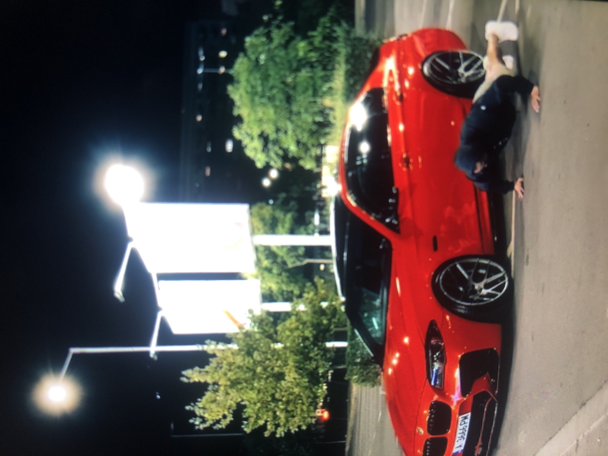 A red BMW with plate number "A-3666-PM" is parked on pavement. An individual wearing white sneakers, light-colored pants, and a dark jacket appears to be inspecting the car. Streetlights and trees are visible in the background, along with what appears to be a sign. The scene is at night. The location cannot be determined as Lhokseumawe.