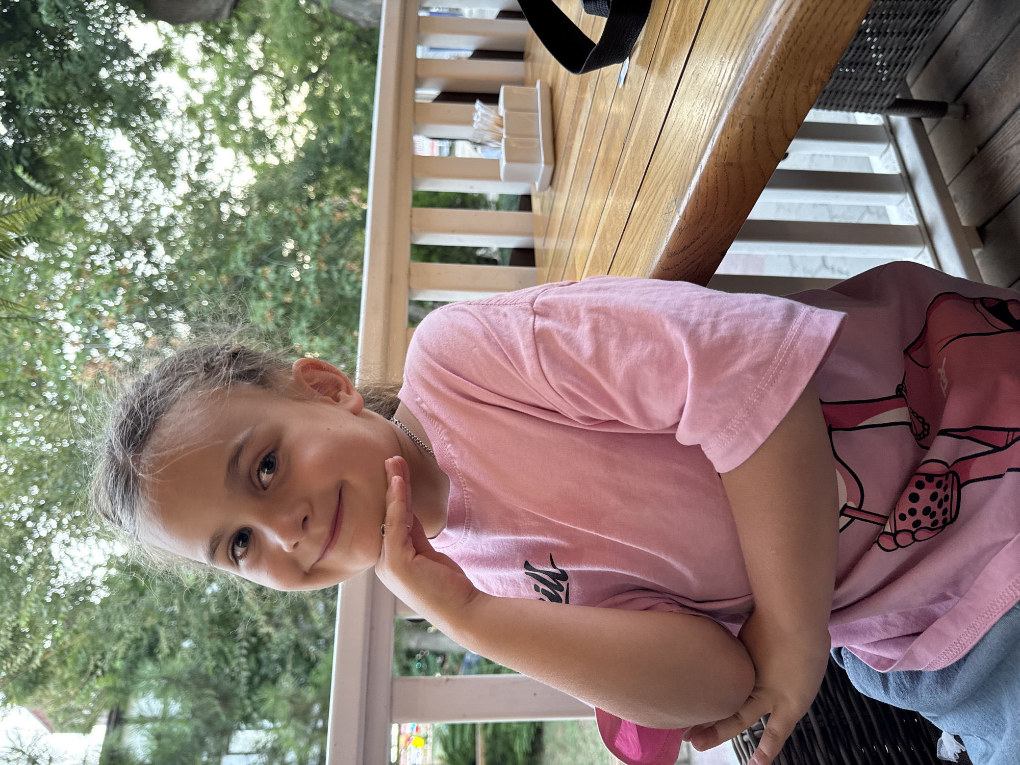 A young girl with blonde hair pulled back in a bun and bangs, is seated at a wooden table on a covered patio. She is wearing a pink t-shirt with a graphic print and blue jeans. Her left arm is bent, and her hand is resting on her chin, giving her a thoughtful expression. The table is set with a white container of toothpicks and a black strap is visible to the right. In the background, a white railing separates the patio from an area with trees and foliage. The setting appears to be an outdoor cafe or restaurant. The image was taken in Yevpatoriya, Ukraine.
