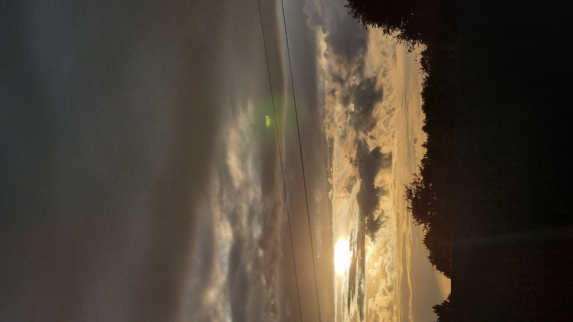 The image shows a cloudy sky with the sun setting or rising, casting a warm glow through breaks in the clouds. Two dark, thin wires are visible horizontally across the sky. Silhouetted trees form a dark line along the horizon, with a darker foreground that appears to be a field or open space. The location is identified as Vysokoe, Russia.