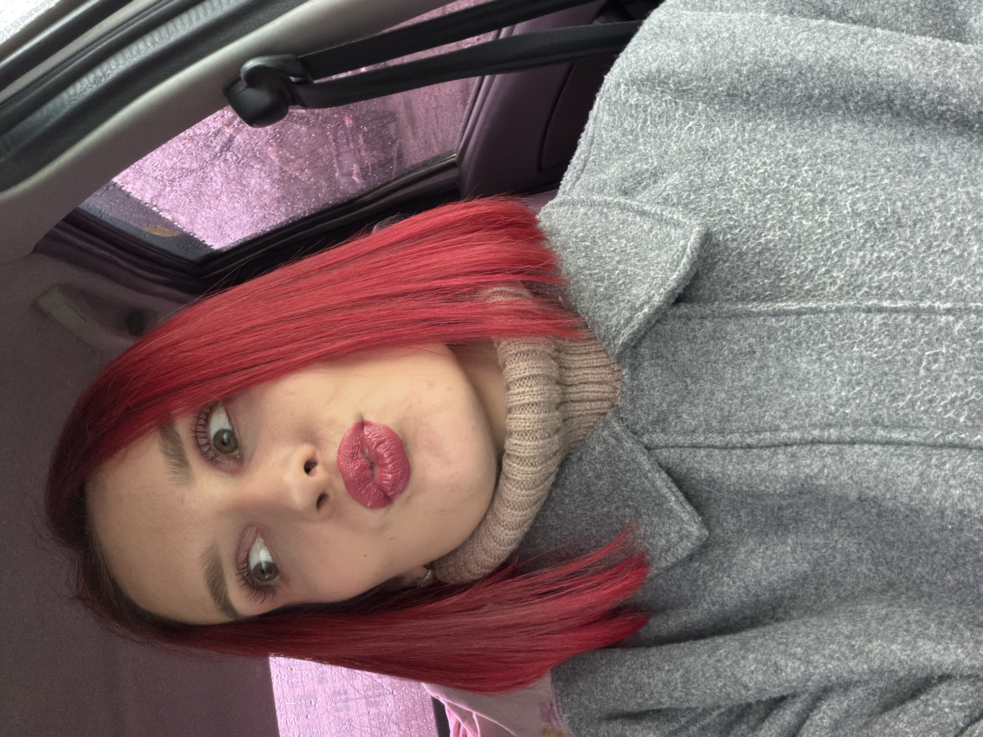 This media file is a selfie of a young woman, captured with the image rotated 90 degrees counter-clockwise, taken inside a car in Chaikovsky, Russia. The woman features striking, long, straight, vibrant red hair. She is wearing a warm, light gray coat, distinguished by its textured, possibly wool or fleece, fabric, layered over a beige, ribbed turtleneck sweater. Her lips are puckered in a playful "kissing" pose, accentuated by a matte pinkish-fuchsia lipstick. Her expressive eyes, framed by defined eyebrows and dark mascara, gaze directly at the camera.
The setting reveals the interior of a car, with parts of the headliner and a sunroof mechanism visible. Through the car's sunroof, a blurred, textured expanse of purplish-pink is seen, suggesting an overcast sky or a reflection during what appears to be a rainy day, as subtle water droplets are evident on the glass. The lighting is diffused, consistent with an outdoor scene under cloudy conditions. No text is visible in the image. The overall mood conveyed is casual and expressive.