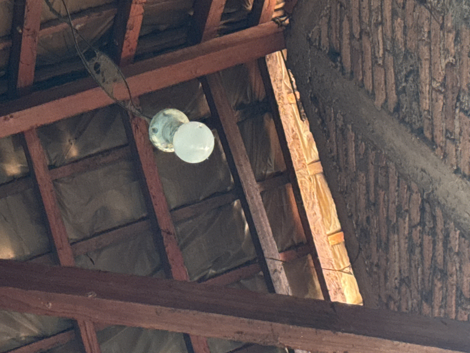 An old, lit light bulb hangs from a wire amidst the exposed wooden beams and roofing structure of what appears to be an attic or unfinished building in Slahung, Indonesia. The light bulb, a translucent white, illuminates the surrounding area with a soft glow. The wooden beams, weathered and reddish-brown, form a diagonal pattern against a backdrop of grey insulation material. Below the beams, a section of brickwork is visible, partially covered by a layer of rough, grey mortar or cement. The overall scene suggests a rustic or aged interior, possibly in a rural or less developed area of Slahung.