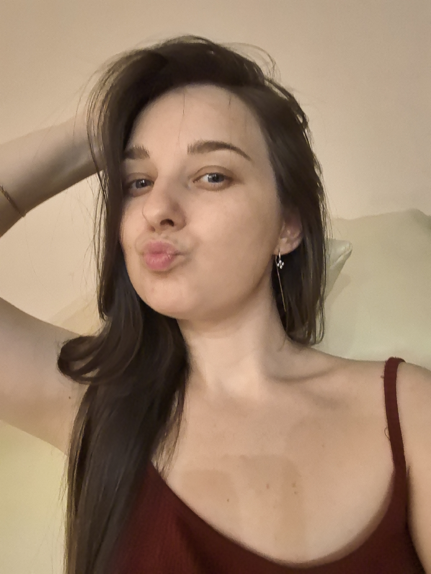 A woman with long, dark brown hair is posing for a selfie. She is wearing a red tank top. She has one hand raised to her head, seemingly adjusting her hair. Her lips are pursed in a kissy face. The background is a plain, light-colored wall. It is not possible to determine the location of the image.