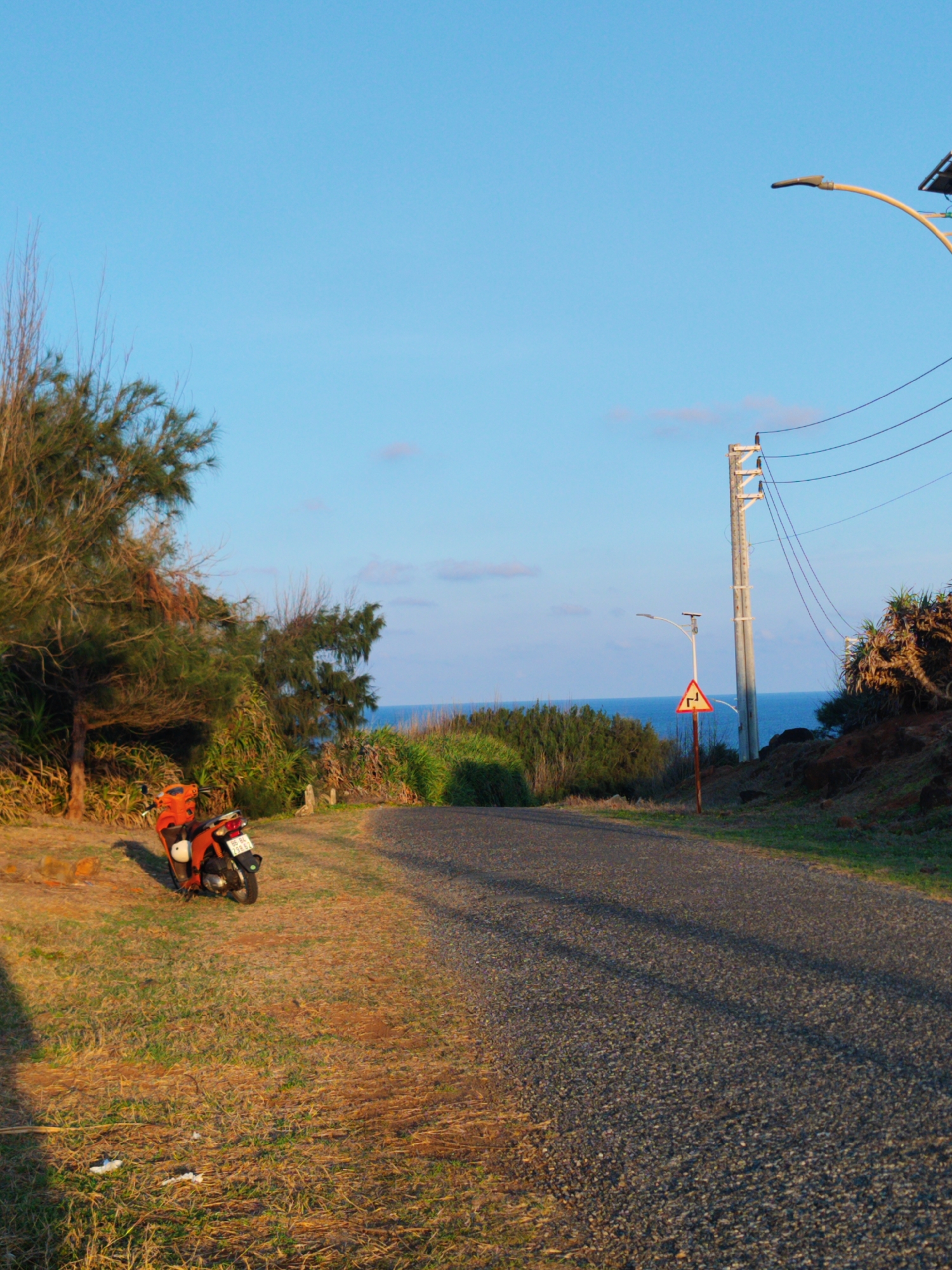 An asphalt road curves along the coast on Phú Quý Island, Phú Quý Special Administrative Region, Vietnam. A moped is parked on the grass along the roadside to the left. A warning sign sits on the right side of the road. Utility poles are also visible. The sea is in the background. The sky is blue and clear.