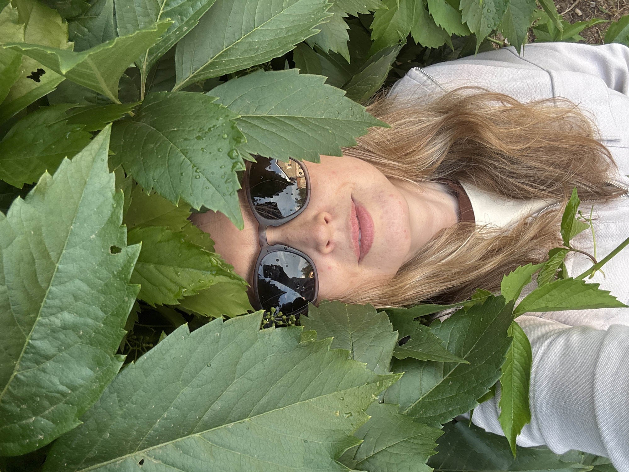 A woman wearing sunglasses and a light-colored hooded jacket peeks out from behind lush green foliage. Her blonde hair is visible, and her lips are slightly parted. The sunglasses reflect a building with a tiled roof and a cloudy sky. The foliage appears to be growing outdoors, possibly in a park or garden setting within the city of Chaikovsky, Russia. The leaves are large and serrated, with some appearing to be damp from recent rain. The overall impression is one of a casual, outdoor portrait set against a natural backdrop.
