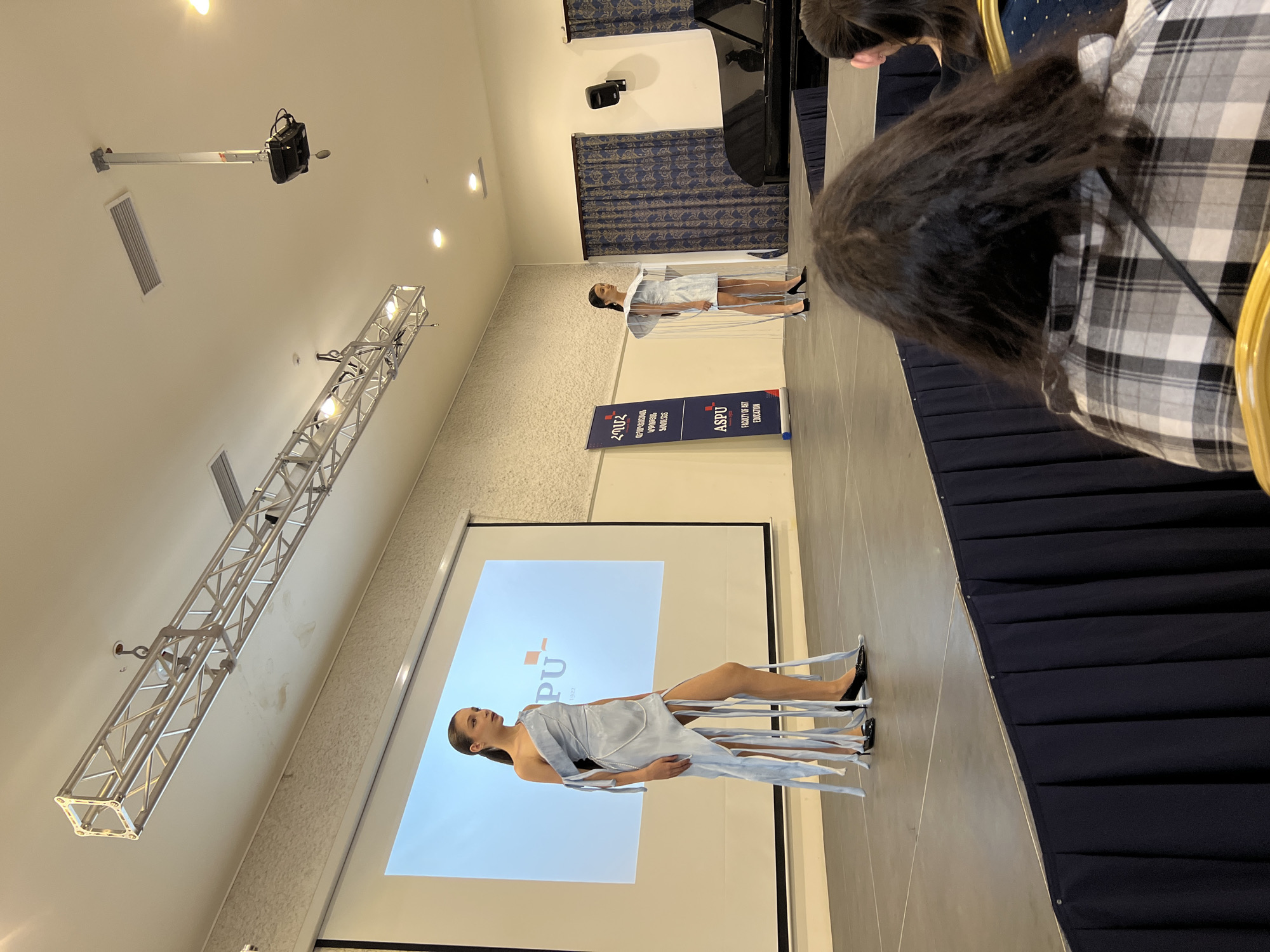 The image shows a fashion show taking place in a conference hall with a high ceiling. One model is on the runway, wearing a light-blue dress with draped ribbons or fabric strips. The show is being recorded or projected onto a large screen behind her, showcasing a stylized “ASPU” logo. The background suggests a conference room, complete with dark blue curtains. Another model is farther back on the runway. The setting appears to be Yerevan, Armenia, based on the logo displayed on the banner: "Armenian State Pedagogical University after Khachatur Abovyan". A stage lighting truss hangs from the ceiling above. There are people in the foreground watching the show.