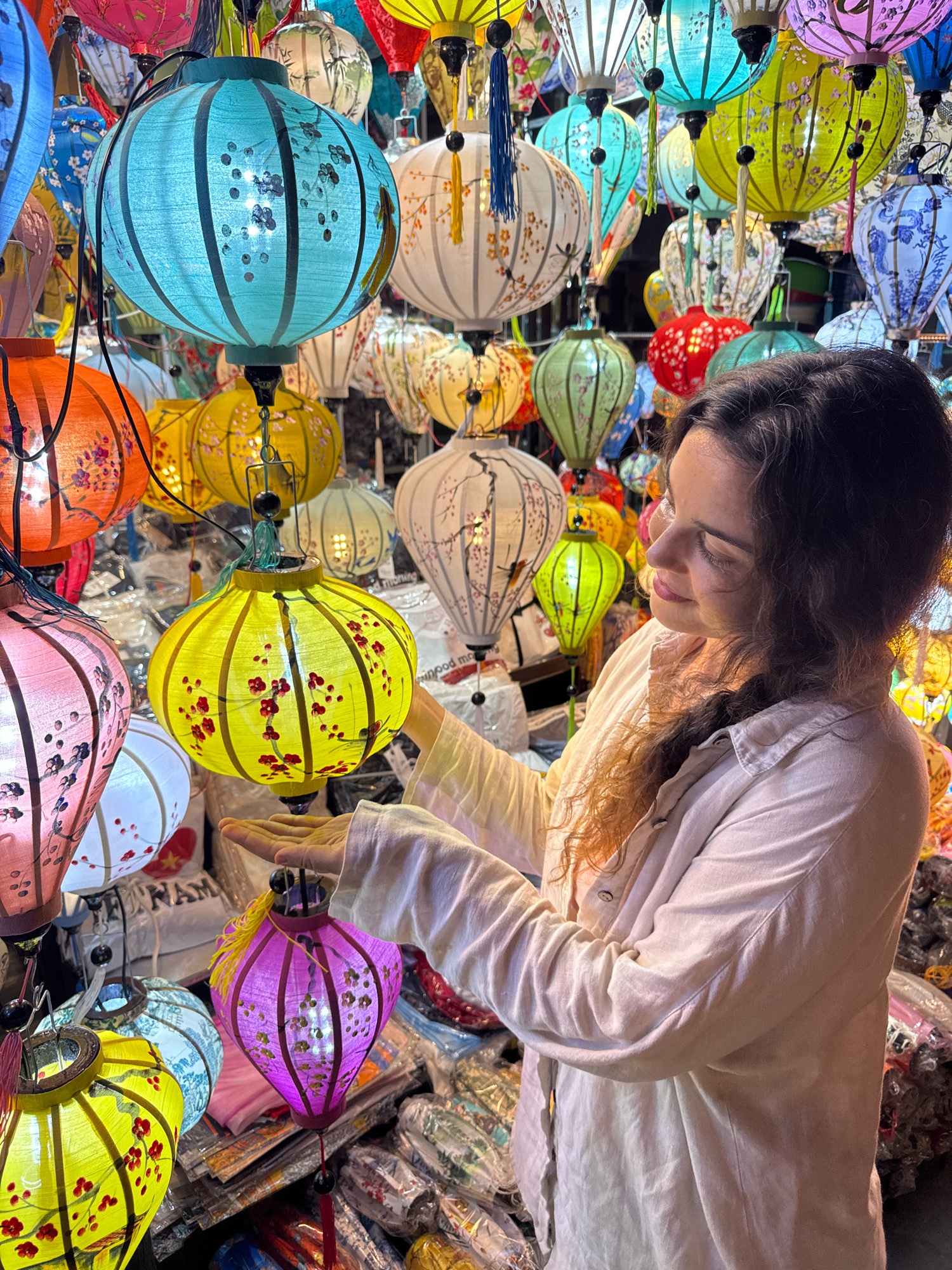 A young woman with long, curly brown hair, wearing a light pink button-up shirt, stands amidst a vibrant display of colorful, patterned silk lanterns in Hoi An, Vietnam. The lanterns, in shades of turquoise, yellow, pink, orange, green, and blue, hang at varying heights, creating a festive and enchanting atmosphere. Some lanterns are decorated with floral patterns, while others feature intricate imagery. The woman is captured in a moment of engagement, with her right hand extended towards a bright yellow lantern adorned with red flowers, her gaze directed towards it with a gentle smile. The background is filled with more lanterns and the soft glow of their internal lights, suggesting a bustling market or shop. The scene is illuminated by the ambient light of the lanterns and perhaps the surrounding environment.