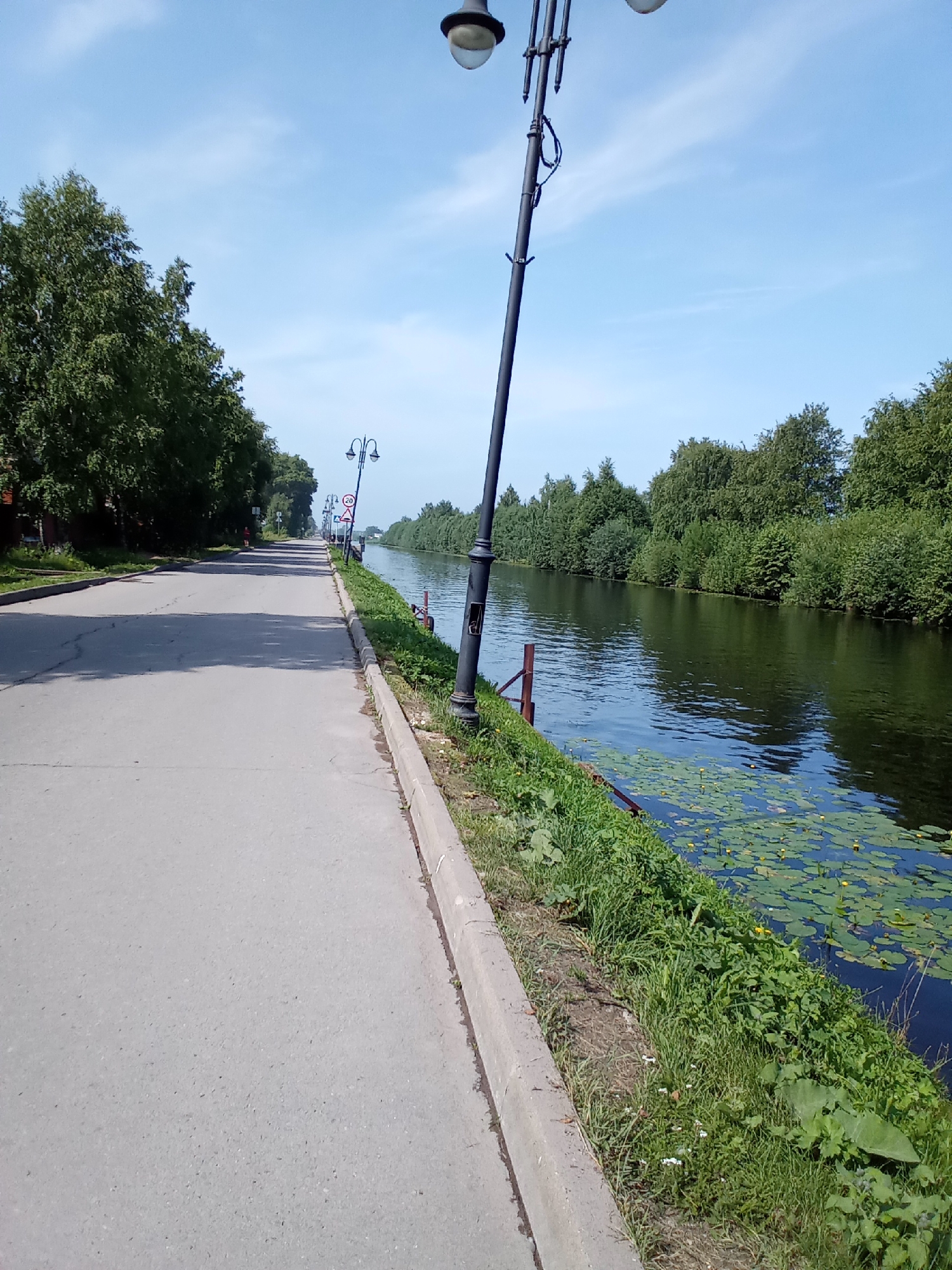 The image depicts a scenic canal lined with trees and a paved walkway in Belozersk, Russia. A street lamp with multiple lights stands prominently on the right side of the frame, casting a shadow on the walkway. The canal, filled with dark water, is partially covered with lily pads and yellow flowers. The walkway is bordered by a concrete curb and green grass. On the left side of the image, lush green trees line the street, with a few buildings visible in the distance. The sky is clear and blue with scattered white clouds. The scene appears peaceful and serene, suggesting a pleasant day.