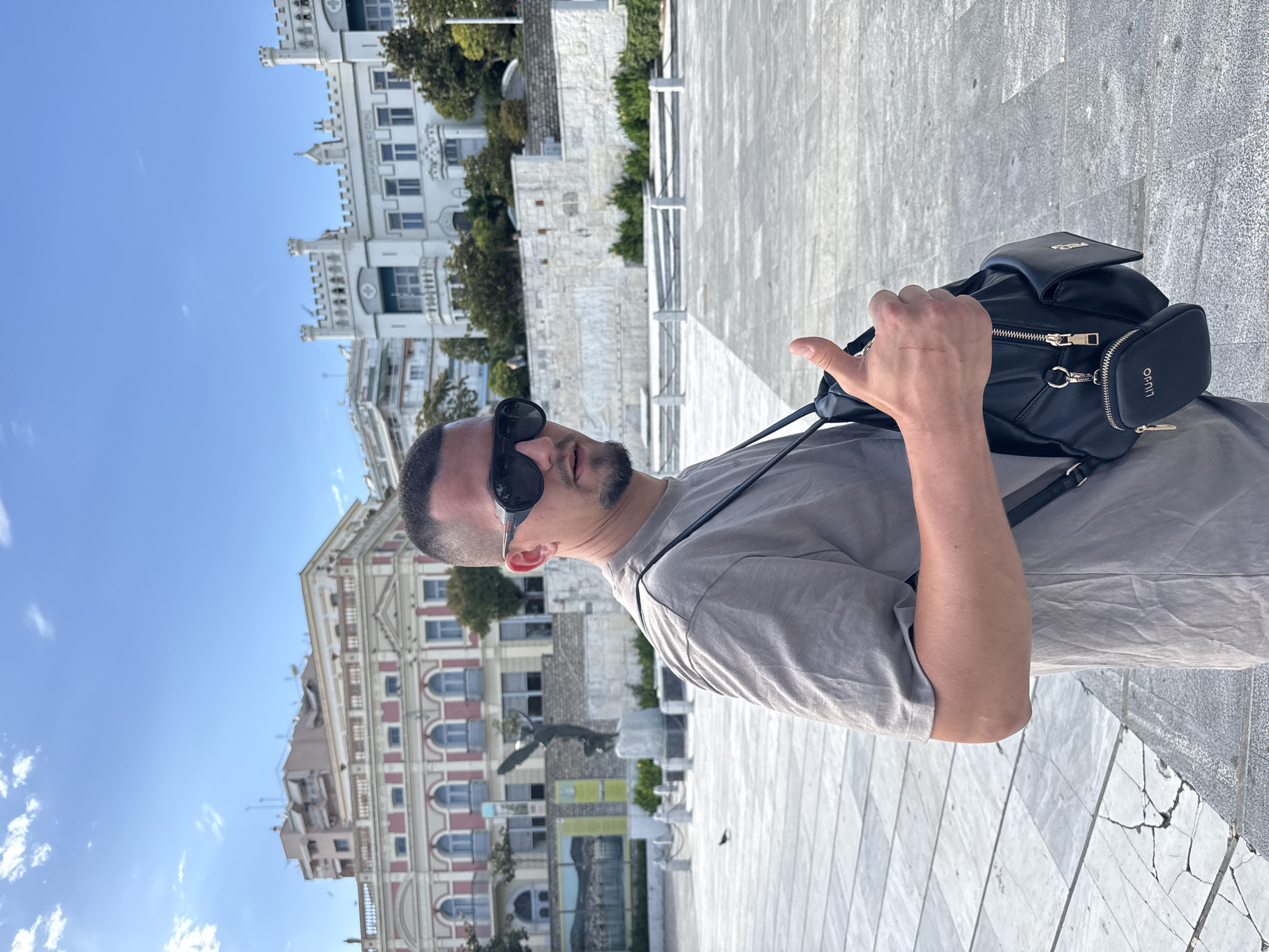 A medium shot captures a young man, facing right and looking towards the camera, in an outdoor urban setting under a bright, partly cloudy sky. He has a buzz cut, a short beard, and wears dark, oversized sunglasses. His left arm is bent, with his thumb extended upwards in a "thumbs up" gesture. He is carrying a black backpack over his right shoulder, with a small black zippered pouch attached to the front by a metal clasp. The pouch has the word "LIUJO" printed on it in white. The man is wearing a plain, light brown t-shirt.
The background features a paved plaza with a light grey, squared stone pattern. In the mid-ground, a low wall with decorative stonework runs horizontally. Behind the wall, a grey stone statue of a winged figure stands on a pedestal. Further back, two distinct buildings are visible. The building to the left is a large, ornate structure with a pink facade, arched windows, and a cream-colored upper story. The building to the right is a white, castle-like structure with crenellated battlements and multiple windows. Trees and some foliage are scattered throughout the mid and background. The ground shows some cracks in the paving stones. A single black bird is visible on the plaza floor in the mid-left of the frame.