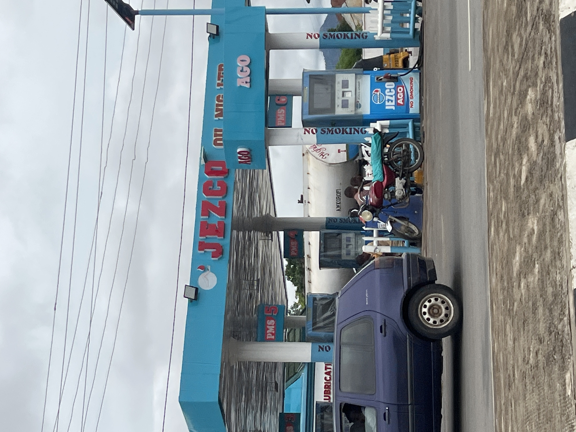 This image captures a JZCO oil and gas station in Jalingo, Nigeria, under an overcast sky. A blue and white canopy covers multiple fuel pumps. The prominent signage reads "JZCO OIL NIG. LTD." and displays "AGO" and "PMS" along with numbers indicating fuel prices (e.g., "PMS 5"). One fuel pump station is branded with "JZCO AGO" and "NO SMOKING" warnings on its sides. A dark purple station wagon is parked on the left, facing away from the camera. To the right, a red motorcycle is parked, and several individuals are present around the fuel pumps, including a person attending to a fuel pump. In the background, a large white fuel tanker is visible, with "AMKOROF" written on its side. The overall environment suggests a functional, working gas station.