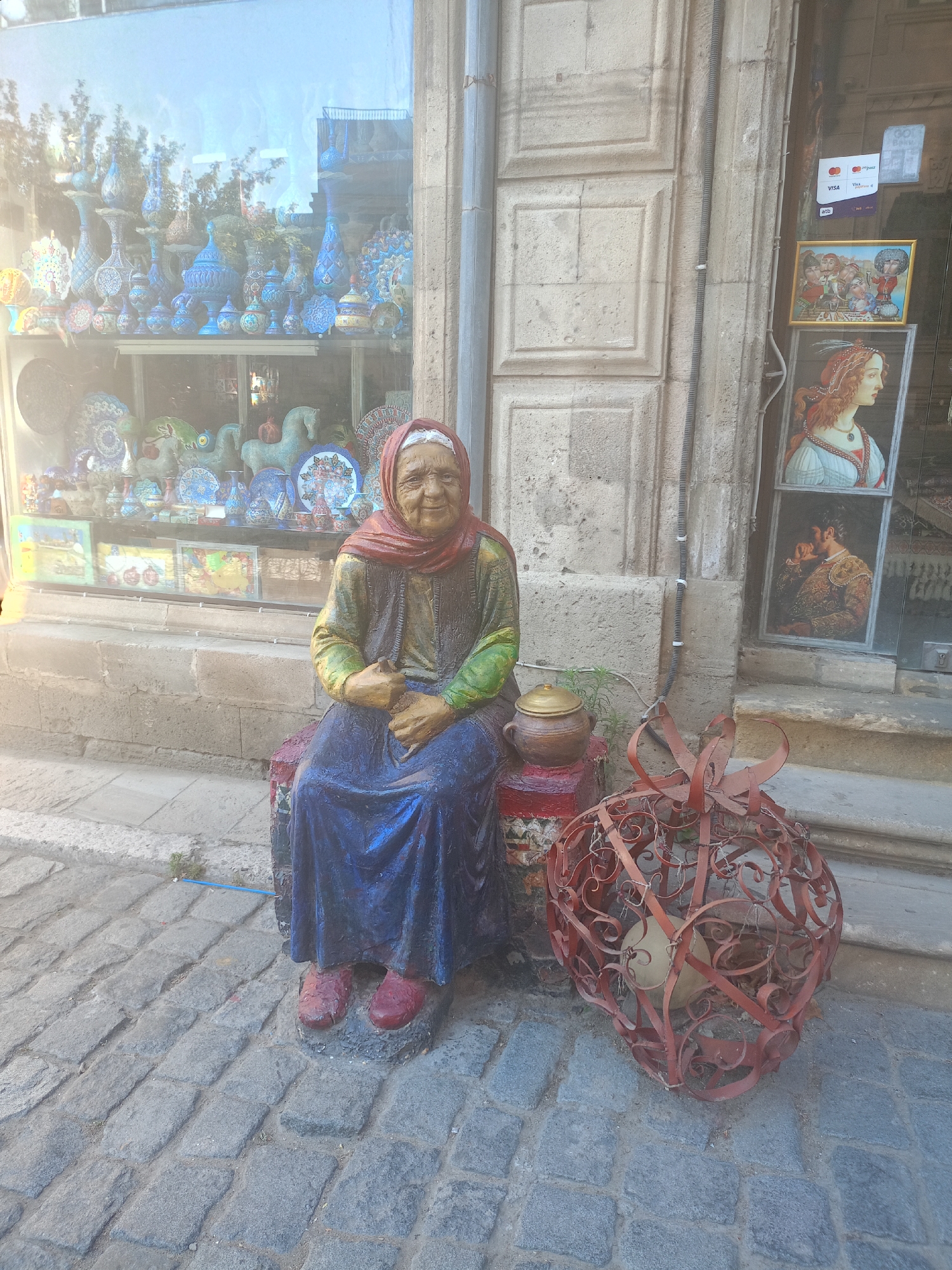 An outdoor, eye-level, medium shot shows a statue of an elderly woman sitting on a small, decorated stool in front of a shop window. The statue, which is life-sized, depicts a woman with a weathered face, wearing a red headscarf, a green and yellow long-sleeved shirt, and a dark blue full-length skirt. Her hands are clasped in her lap, and she is wearing red shoes. To her right, on the cobblestone ground, is a large, ornate, red metal lantern with a light-colored spherical object inside. To the statue's left, a portion of a shop window is visible, displaying an array of colorful pottery, vases, and decorative plates. The shop's facade is made of rough, light-colored stone blocks. The ground is paved with irregular gray cobblestones. In the upper right portion of the image, a framed painting of a woman with red hair and a painted man with a mustache are visible through the shop window, along with payment stickers. The lighting suggests it is daytime, with some glare on the shop window.