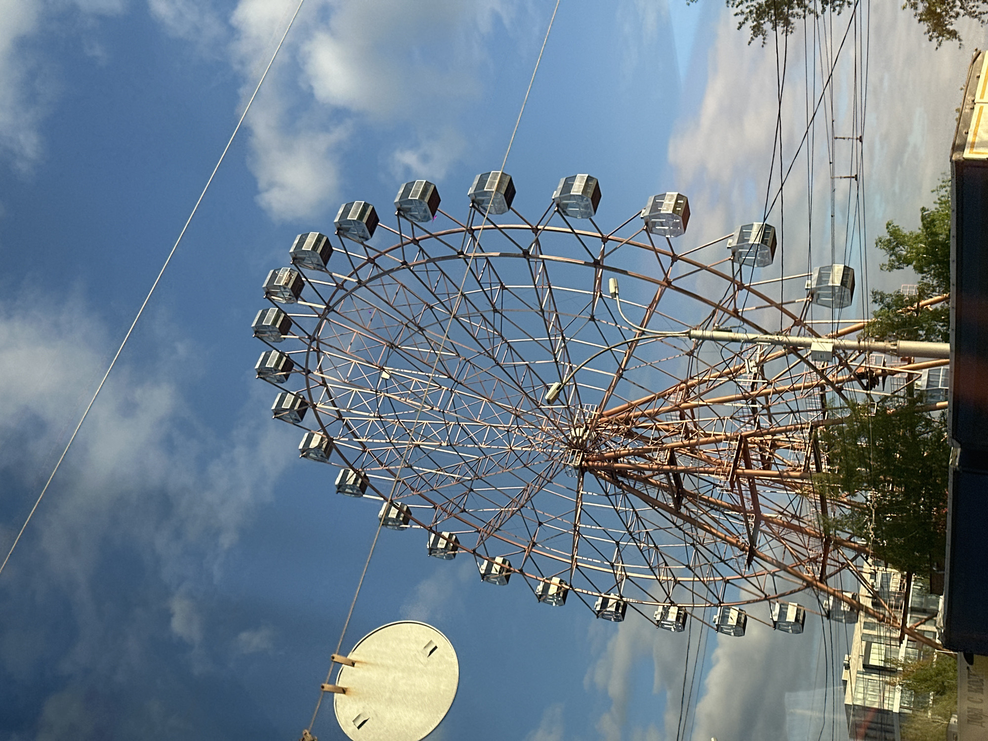 The image shows a large Ferris wheel against a blue sky with scattered clouds. The Ferris wheel has numerous enclosed gondolas, each with a boxy design and light-colored framing. The wheel's structure is made of metal lattice. A partially visible white disc with metal supports is in the lower-left corner. Power lines are visible above the wheel and a fragment of a building and tree branches appear on the right. The location is Novosibirsk, Russia.