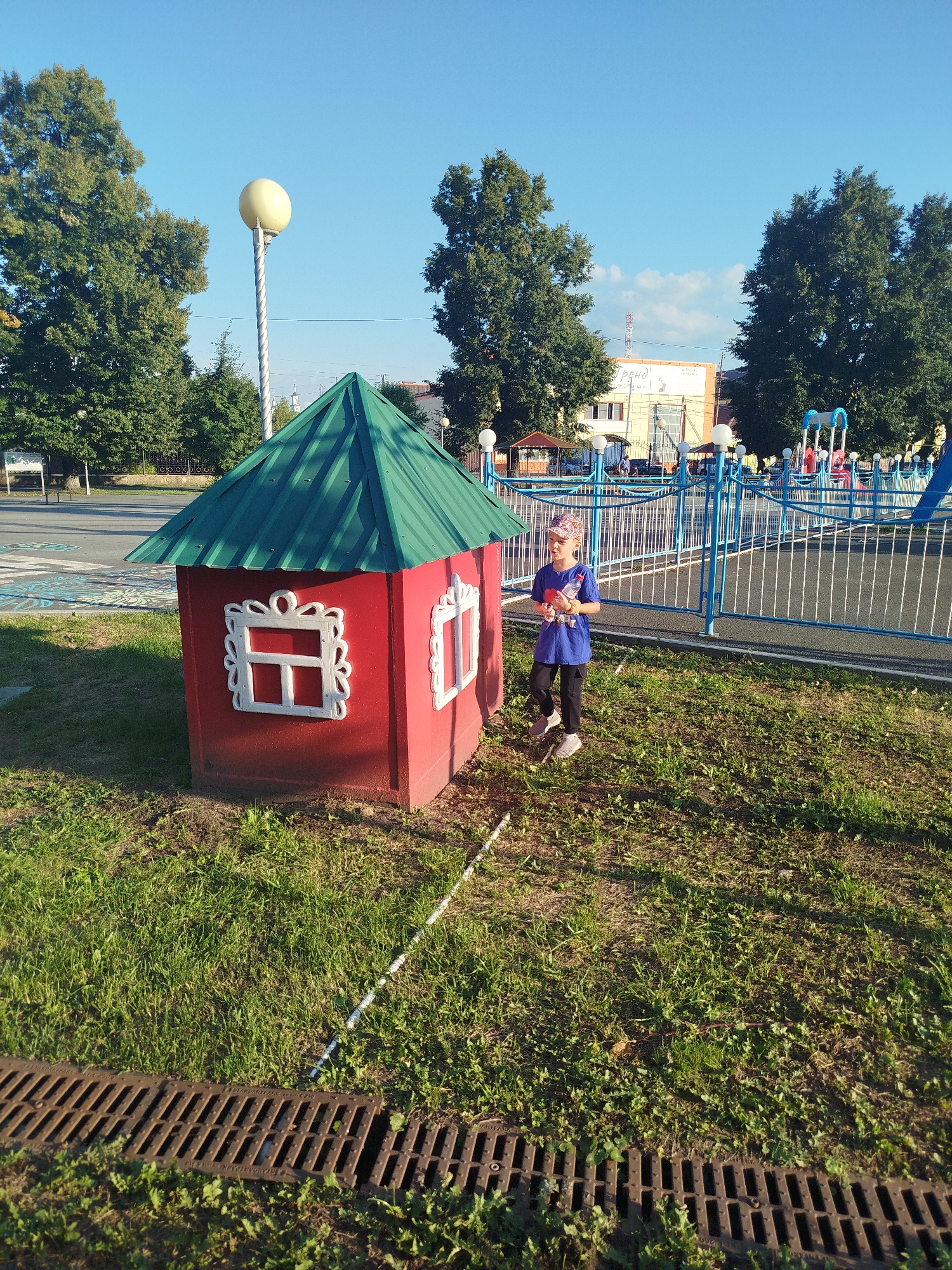 A child wearing a blue t-shirt, dark pants, and a patterned cap stands in a grassy area next to a small, red, toy house with a green roof. The child is holding a red object. Behind the child, a blue metal fence encloses a playground area with slides and swings. Large green trees are visible in the background, along with buildings under a clear blue sky. The scene appears to be a public park or playground in the city of Yalutorovsk, Russia. A metal grate for drainage is visible in the foreground.