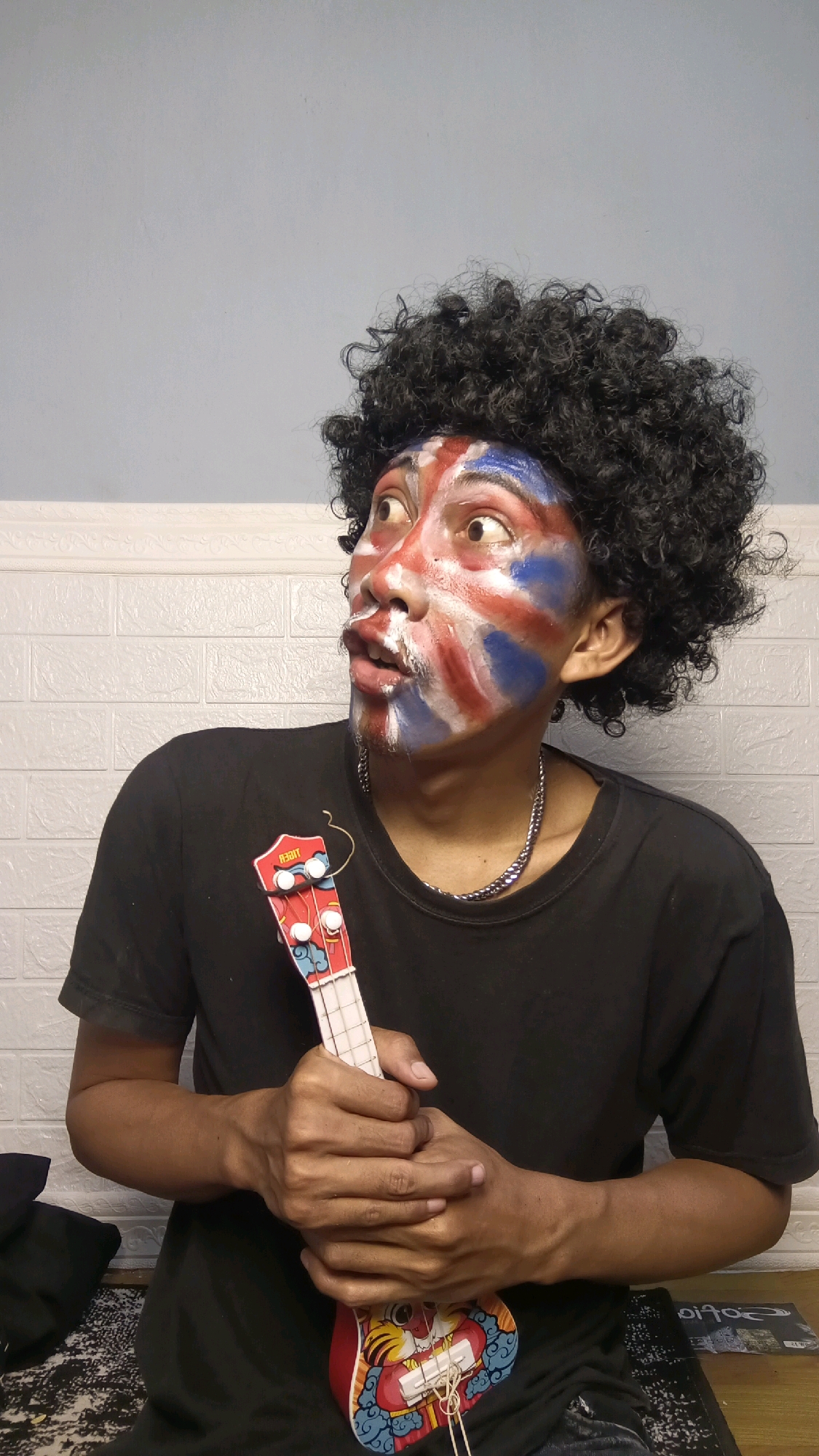 A young man with a curly black wig and face paint resembling the Union Jack flag poses with a red ukulele. He is wearing a black t-shirt and a silver chain necklace. The background consists of a light blue wall above a white brick textured wall. The scene appears to be indoors, possibly in a residential setting in Kebumen, Indonesia.