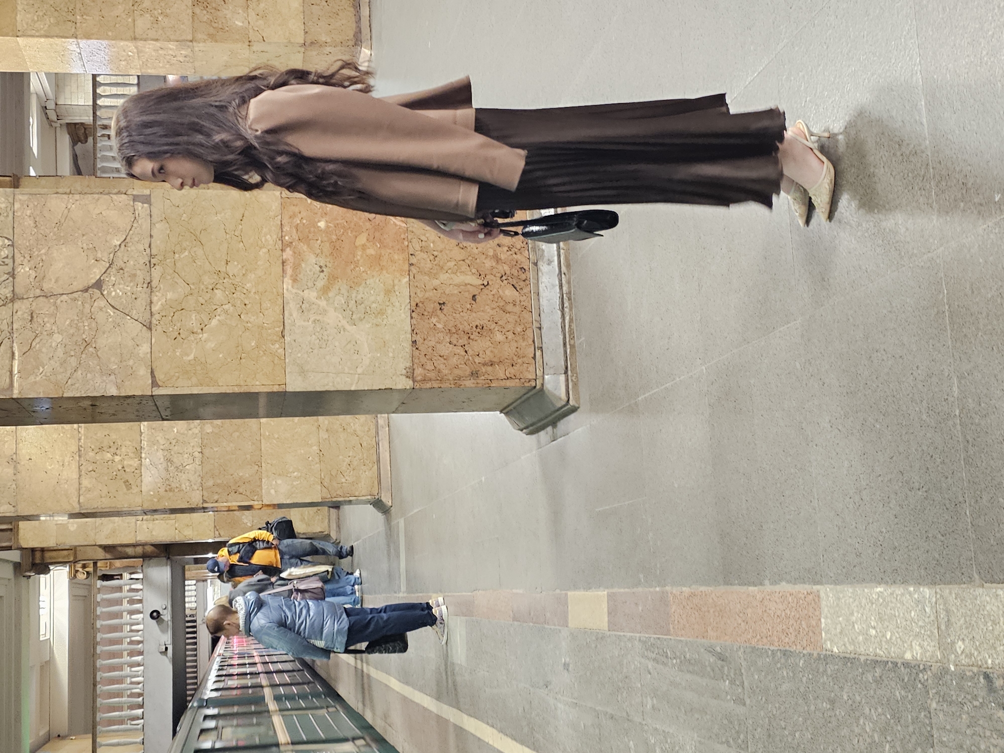 A full shot, rotated 90 degrees, captures a woman in a brown coat and dark skirt holding a black purse. She is walking on a grey tiled floor adjacent to a wall of mottled, light-brown stone slabs separated by silver metal bars. Other people are partially visible to the left of the frame waiting for public transportation. The location is Moscow, Russia.