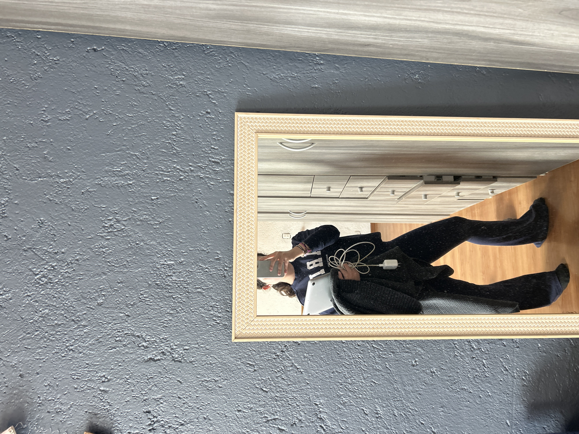A person is seen taking a mirror selfie indoors, with the image rotated 90 degrees counter-clockwise. The individual, reflected in a rectangular mirror, holds a smartphone to capture the picture and a silver laptop. A white charging cable with a power adapter is draped over their arm. They are wearing a dark long-sleeved top with partially visible white lettering "1987" on the front, dark pants or leggings, and dark socks.
The mirror has a light-colored frame featuring a distinct basketweave pattern. The reflection inside the mirror reveals a room with light-colored walls or furniture, possibly a dresser or wardrobe with multiple drawers, and a light wood-colored floor.
The wall outside the mirror is a heavily textured, dark gray-blue color. Above the mirror, a section of horizontally oriented light gray wood paneling or trim is visible. The lighting suggests it is daytime.