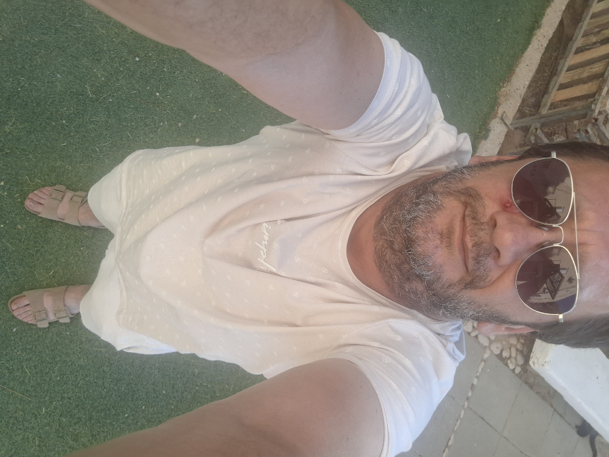 A man is taking a selfie outdoors. He is wearing sunglasses, a light-colored t-shirt, and sandals. He has a beard and appears to have a small injury on his cheek near his eye. The ground is covered in green grass, and there is a wooden fence and paved area in the background. The image is taken from a high angle, looking down at the man. The scene is likely in Yerocham, Israel, given the location context.