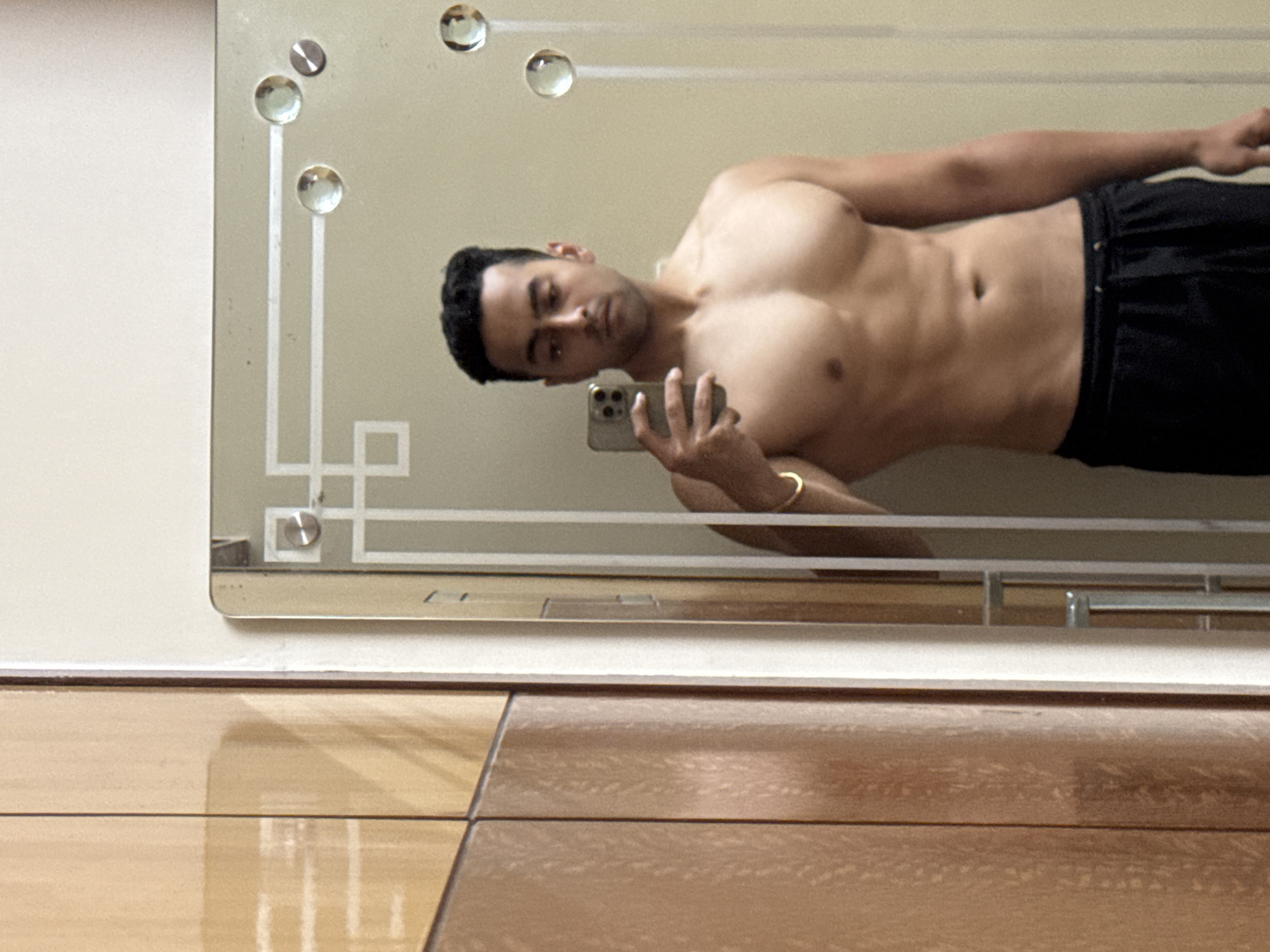 A shirtless man is taking a selfie in a mirror. The man has short dark hair and is wearing dark-colored shorts. He is holding a phone in his left hand, with his right arm extended out to the side. The mirror has decorative elements including small clear spheres and geometric patterns in white. The background is a plain wall, and the floor is tiled. There is no evidence in the image to indicate the city of Kolkata, India.