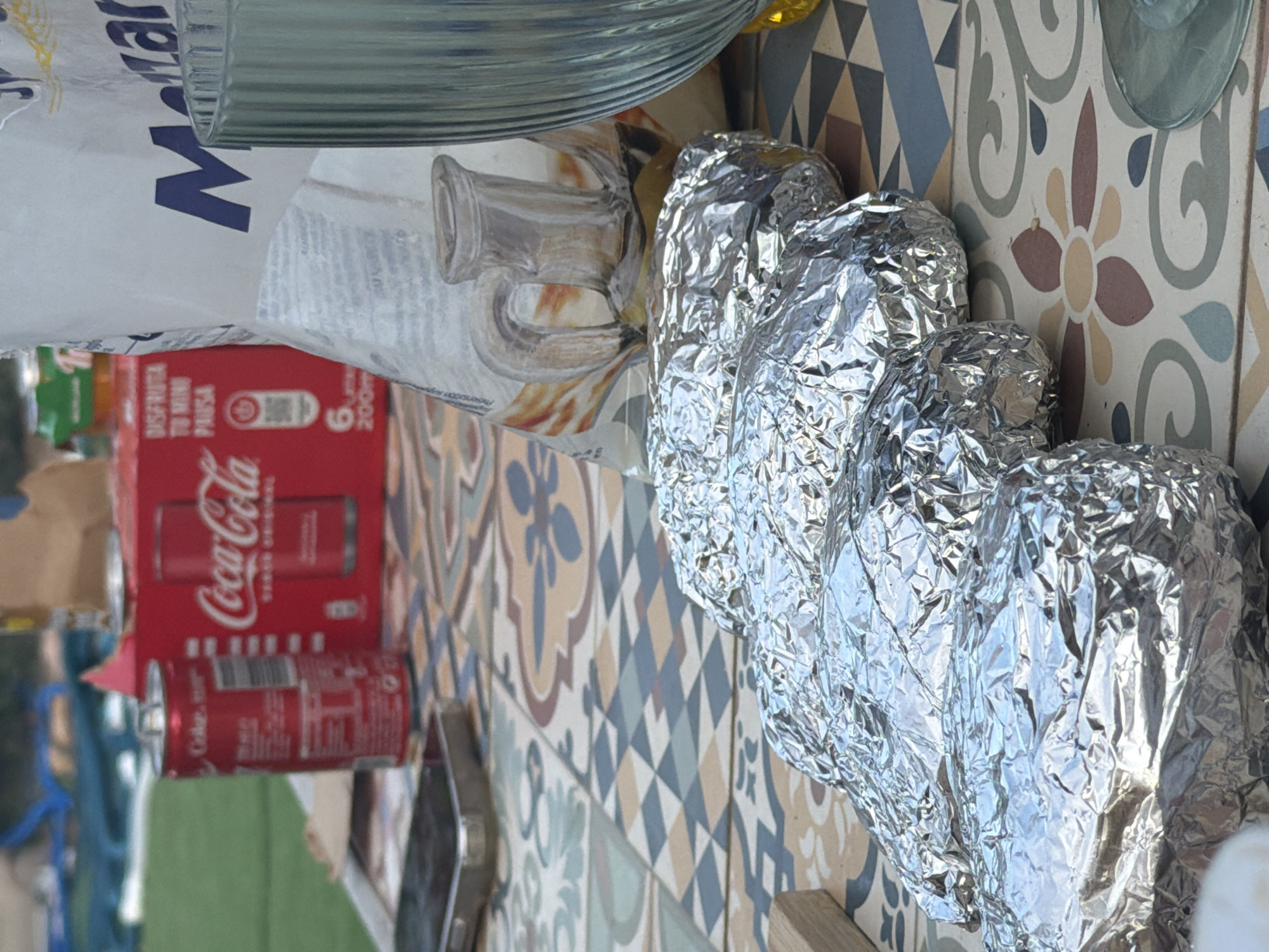 The image shows a close-up view of a table laden with food and drinks, suggesting an outdoor gathering or picnic. In the foreground, four foil-wrapped items, likely food such as baked potatoes or empanadas, are arranged in a row. The table surface is adorned with a colorful, patterned tile design. Behind the food, a red Coca-Cola carton is visible, along with a single can of Coca-Cola. To the right, a tall, ribbed glass, possibly for a beverage, stands beside a white bag with blue lettering that reads "Malt". A portion of a smartphone is also visible on the table. The background is blurred, showing hints of greenery, suggesting an outdoor setting. The lighting indicates it is daytime, with natural light illuminating the scene. There are no people visible in the frame.