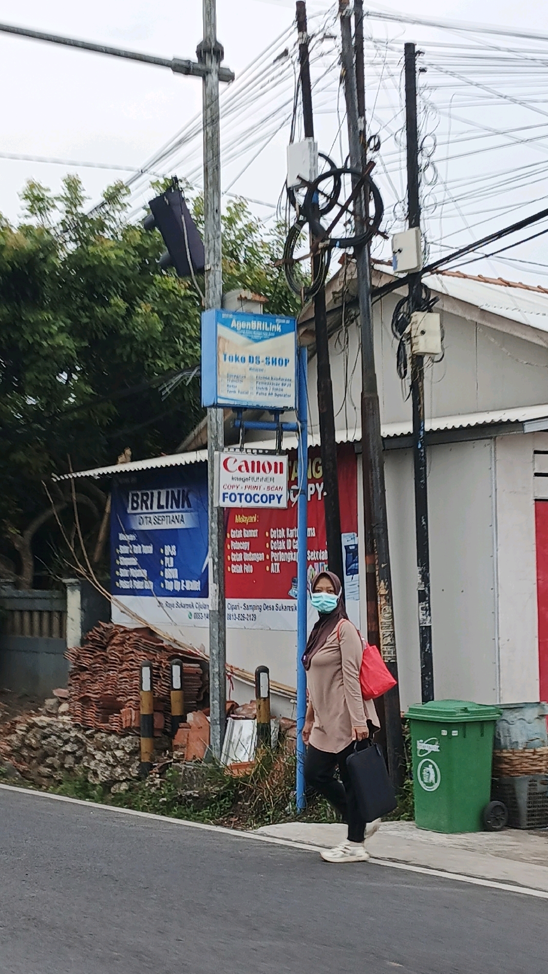 This is a medium shot of a woman walking on the right side of a street in Sukaresik, Indonesia. The woman is wearing a light brown long-sleeved shirt, black pants, a green hijab, and white sneakers. She is carrying a black tote bag in her right hand and a red bag over her left shoulder. She is facing towards the left of the frame and appears to be walking away from the camera.
To the left of the woman are several utility poles with many wires. One pole has a blue sign that says "AgenBRILink" and "Toko DS-SHOP." Below that is a white sign with a red Canon logo and the words "ImageRunner," "Copy-Print-Scan," and "FOTOCOPY." Further to the left is a blue sign that says "BRI LINK DITA SEPTIANA."
Behind the woman and the utility poles is a building with a white exterior and a tiled roof. Next to the building, on the right side of the frame, is a large green trash bin.
The street in the foreground is paved, and there is a sidewalk on the right. On the left side of the street, there is a pile of red tiles and some debris. The overall atmosphere of the image is somewhat cluttered due to the utility poles and wires. The sky is overcast.
