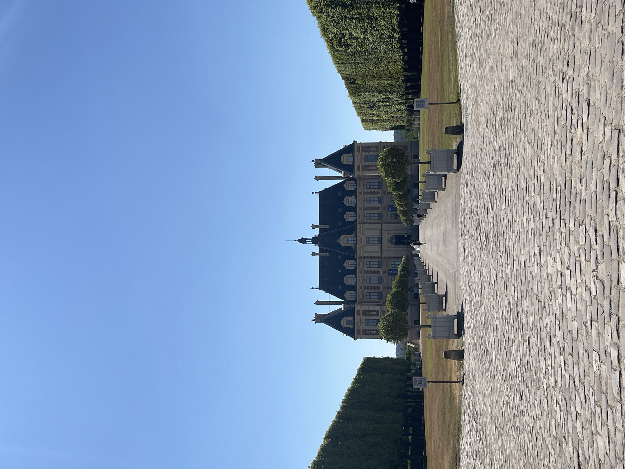 The image depicts the Château de Sceaux, a grand French manor house located in the Parc de Sceaux, a public park in the city of Sceaux, France. The château, a symmetrical building with a central dome and multiple gabled roofs, is framed by perfectly manicured, tall, green hedges on either side. A long, cobblestone pathway leads directly to the château's entrance. Flanking the pathway are rows of potted trees, spaced evenly and creating a sense of formal order. The ground is a mixture of dry grass and cultivated areas. The sky is a clear, cloudless blue. Two figures, appearing to be dressed in white, are visible in the distance, walking along the pathway towards the château. The overall scene suggests a sunny day and a well-maintained, historic estate.