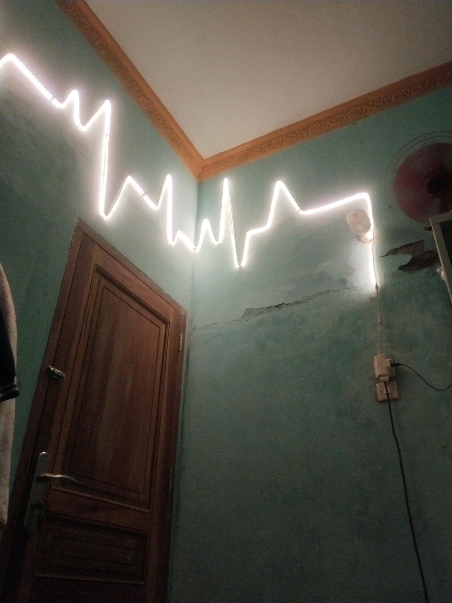 The image depicts a dimly lit room with teal-colored walls and a white ceiling. A decorative wooden door stands on the left side of the frame. A bright white LED light strip, shaped like a cardiograph, is attached to the upper portion of the wall and ceiling, extending across the room. There is a light switch and electrical outlet on the wall to the right. A partially visible red fan is mounted on the ceiling in the upper right corner. The wall appears to have some peeling paint and cracks. The scene is likely an interior space in a residential dwelling in Petanjungan, Indonesia.