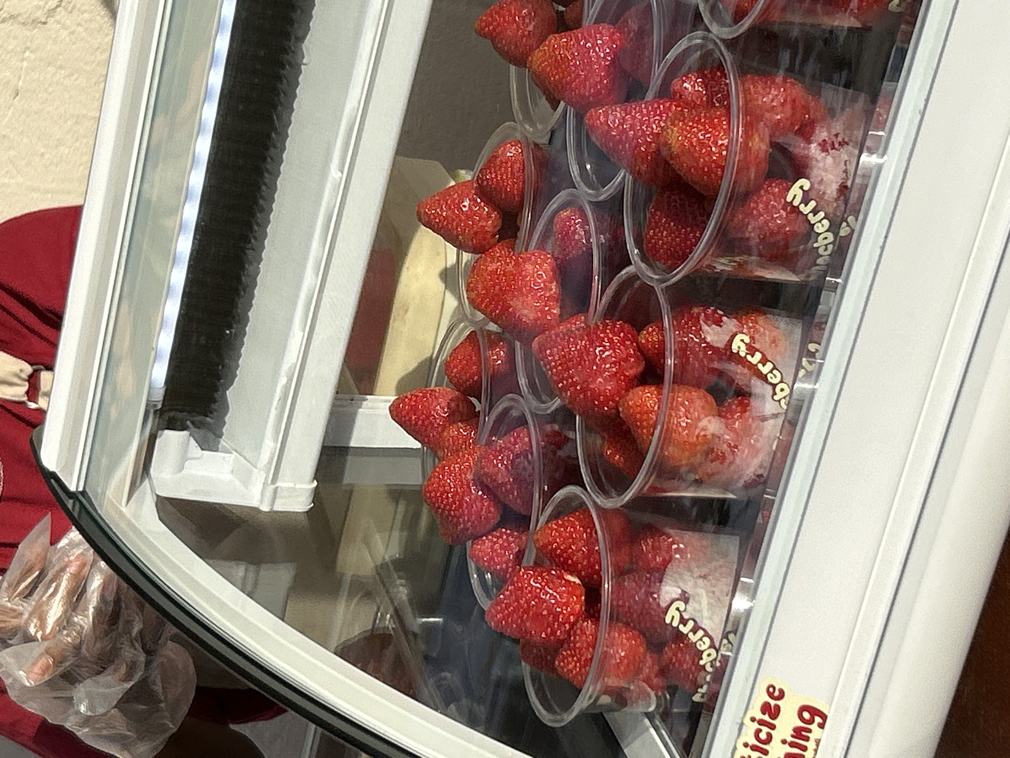 A close-up, eye-level shot shows a display case filled with individual cups of fresh strawberries. The cups are clear plastic and are packed with bright red strawberries. Some cups have a label that says "berry". In the background, a person wearing gloves is seen preparing more strawberries. The display case has a white frame and a glass front. The lighting in the case is illuminated by strip lights. The overall impression is of a fresh fruit stand. The image appears to be taken at a fruit stand in the city of North Kuta, Indonesia.