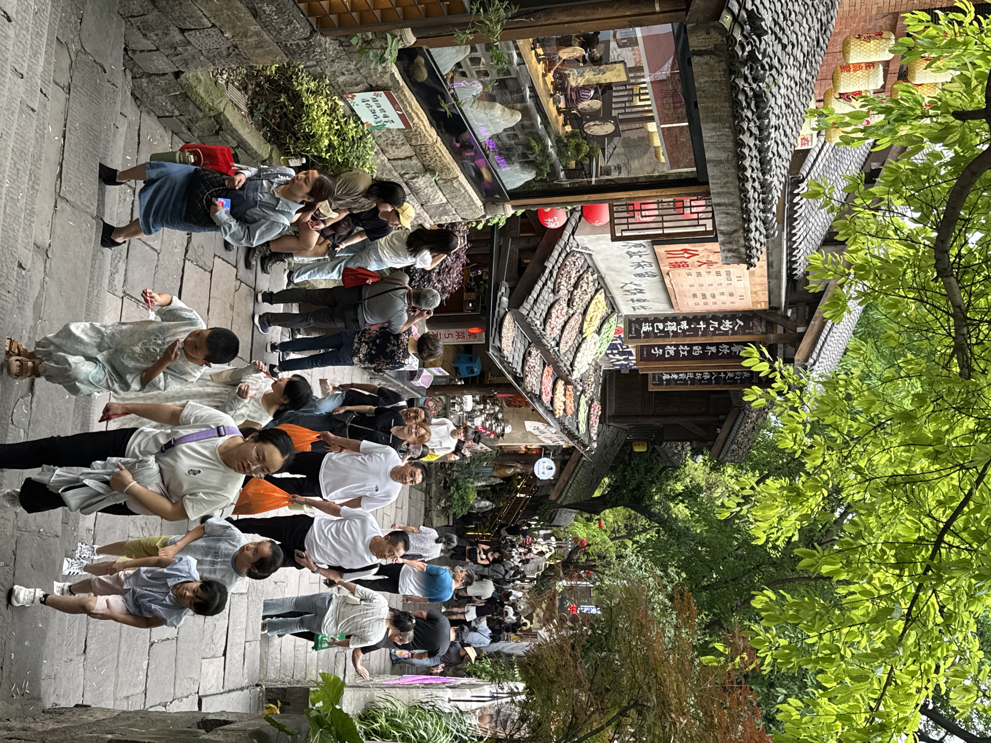 The image shows a bustling street scene in Yuzhong District, China. The narrow stone-paved street is crowded with pedestrians of varying ages, dressed in casual clothing. Many are looking at or walking past shops and stalls lining the street. Overhead, traditional Chinese-style architecture is visible, with tiled roofs and signage. There are trees along the street, and their foliage obscures some of the buildings. The overall environment suggests a tourist area or a popular market street.