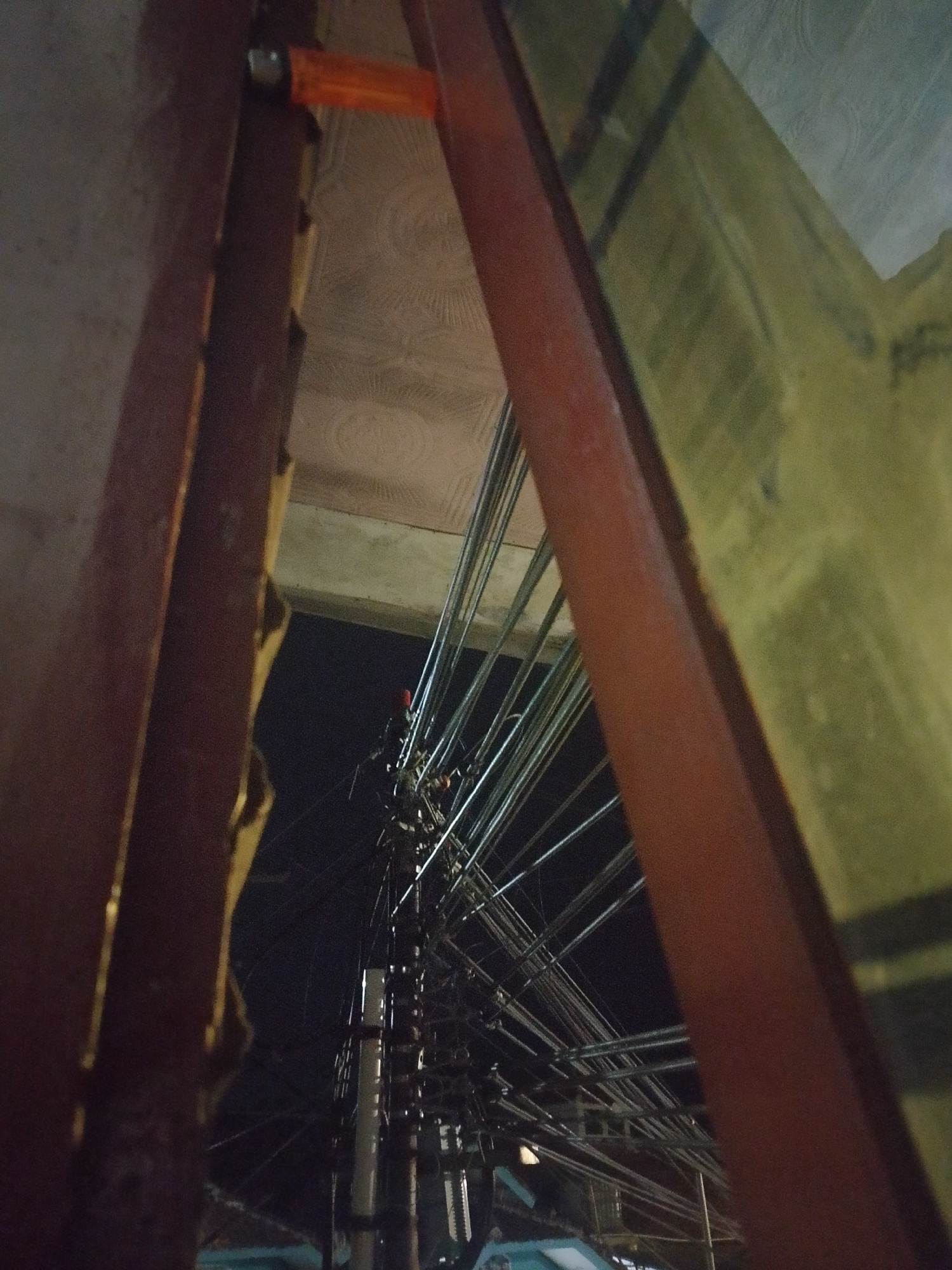 The image is an upward-facing, low-light photograph of an urban environment, likely taken at night. Dominating the foreground are two prominent, reddish-brown structural beams, possibly part of a building or scaffolding, which create a framing effect. Between these beams, a dense network of electrical wires and cables stretches across the frame, suggesting a densely populated area with extensive utility infrastructure. A tall utility pole with multiple crossarms and insulators is visible in the mid-ground, further illustrating the complex wiring. The background is dark, indicating nighttime, but a faint, textured ceiling or overhead structure is visible in the upper portion of the image, possibly the underside of a roof or balcony. A small, orange object, resembling a lighter, is attached to the top of the left structural beam, near the edge of the frame. The overall impression is of an unkempt urban setting with a focus on the utilitarian elements of the city. The location is identified as Mukapayung, Indonesia, suggesting this scene is representative of the visual landscape in that specific locale.