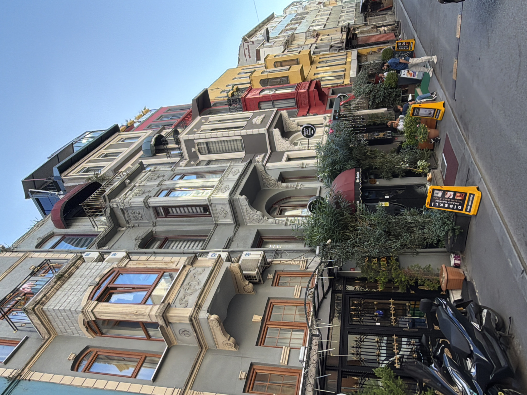 The image depicts a street scene in the Galata neighborhood of Istanbul, Turkey, characterized by its narrow, winding streets and colorful, multi-story buildings. The architecture features a blend of Ottoman and European styles, with ornate balconies, bay windows, and decorative facades. The buildings are painted in various hues, including gray, red, yellow, and blue, creating a vibrant and picturesque atmosphere.
In the foreground, a black motorcycle is parked on the side of the street. A sidewalk cafe with outdoor seating is visible, adorned with lush greenery and string lights. Two men are seated at a table, engaged in conversation, while another man stands nearby, looking at his phone. Yellow A-frame signs with text and images are placed along the sidewalk, likely advertising local businesses or services.
The street itself is paved with cobblestones, and a few people are walking along the sidewalk. The sky is a clear blue, suggesting a pleasant day. The overall impression is one of a charming and lively urban environment, typical of historic districts in Istanbul.