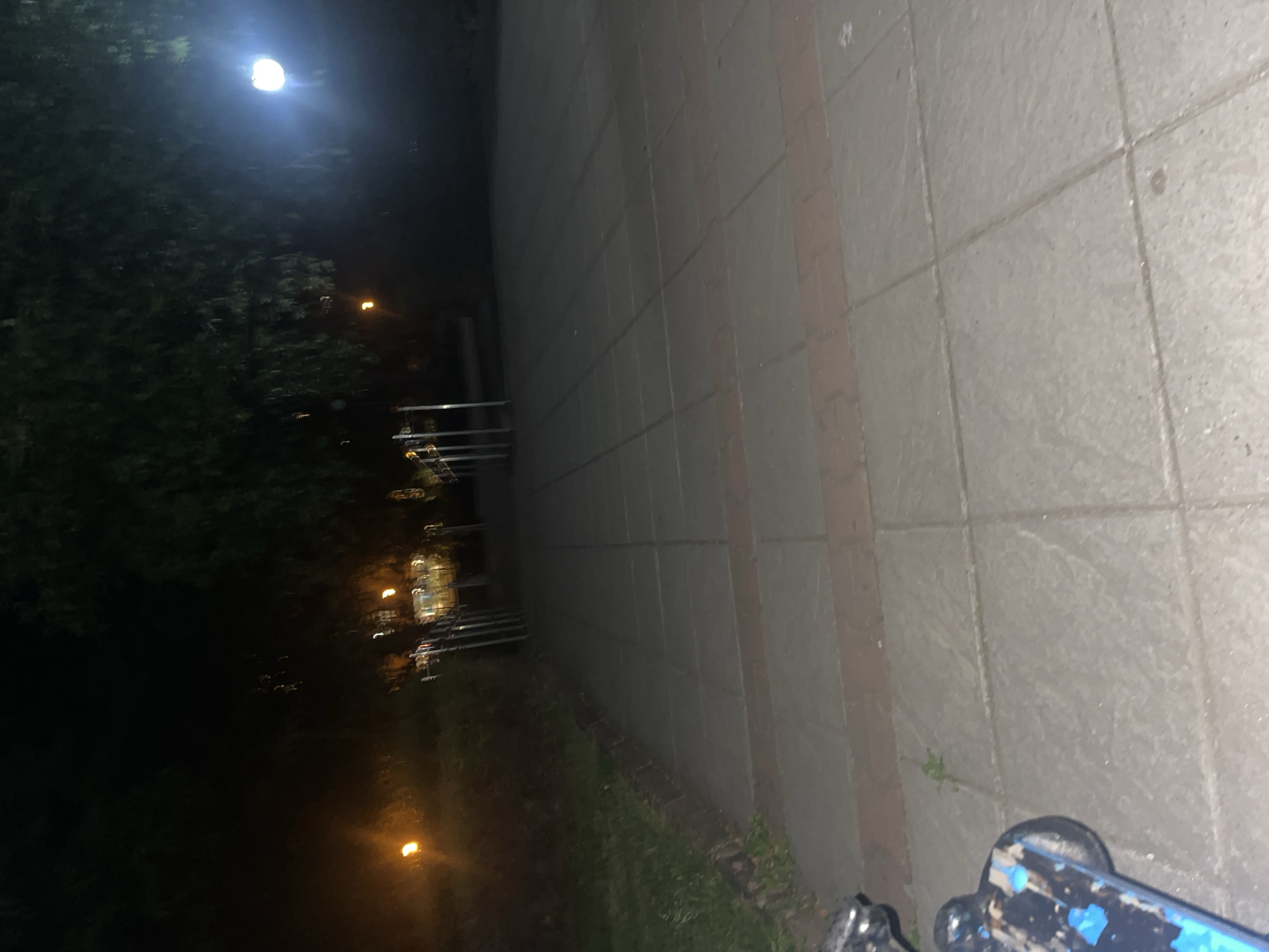 The image depicts an outdoor scene at night in what appears to be a city environment. The right side of the image is dominated by a light-colored, brick or tile wall. Adjacent to the wall is a narrow strip of greenery or grass. In the bottom right corner, part of a blue skateboard is visible. In the distance on the left side, there are trees and street lights. Buildings are suggested by the faint lights and railings visible in the darkness behind the trees. Without additional information, pinpointing the exact location within Blagoevgrad is not possible.