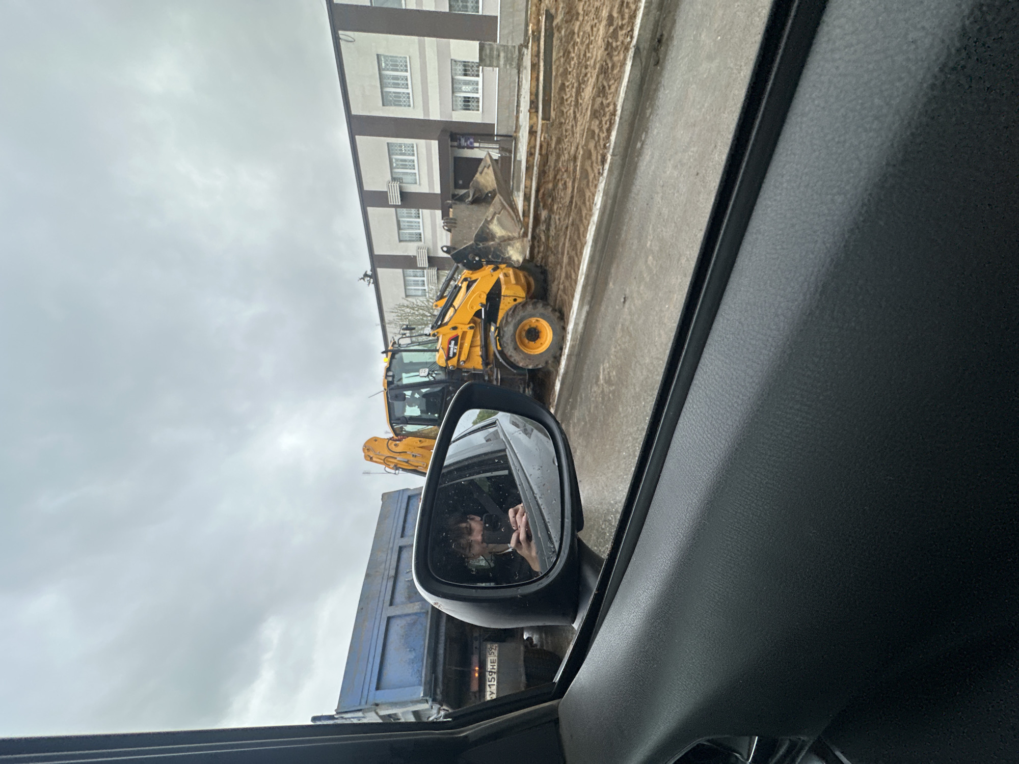 The image shows a view from inside a car. Reflected in the car's side mirror is a blue dump truck followed by a yellow backhoe. Alongside the road, partially visible past the backhoe, is a two-story building. The license plate on the dump truck reads "У 159 НЕ 59". The reflection in the side mirror shows a person holding a phone. The environment suggests an urban or suburban area in Russia, specifically the location of the license plate code being Russia, Chaykovsky. The overcast sky suggests a cloudy day.
