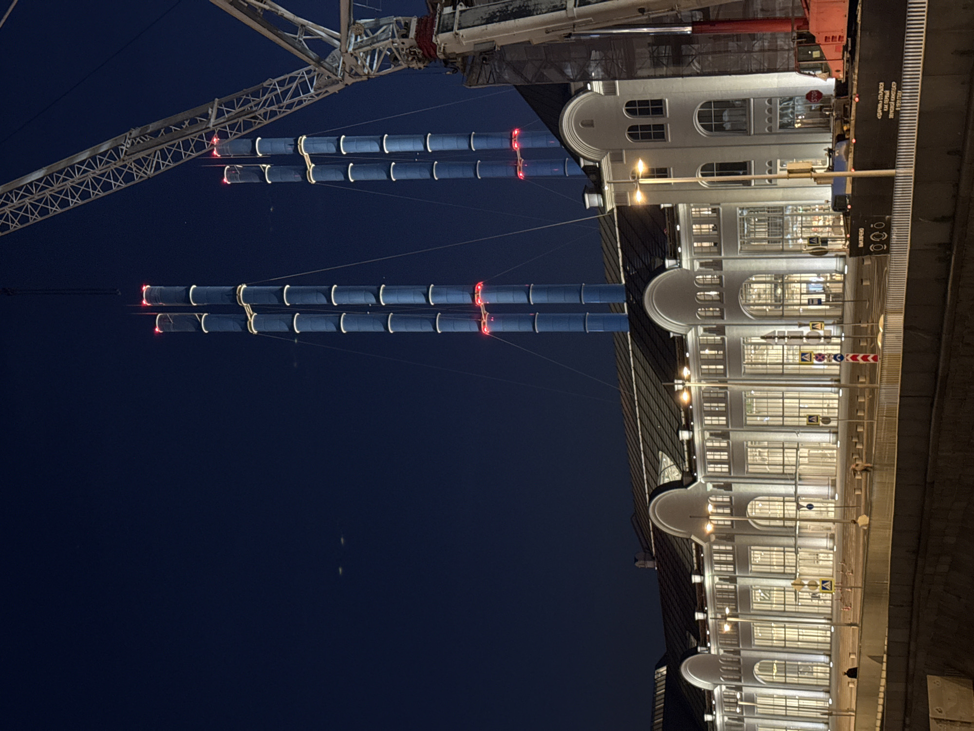 This nighttime image captures a detailed view of the GES-2 House of Culture (formerly Power Station 2) located on Bolotnaya Embankment in Moscow, Russia.
The primary subject is a large, multi-story building with a light-colored facade, featuring numerous tall, arched, and rectangular windows that are brightly illuminated from within. The building exhibits a classic industrial architectural style with repetitive arched doorways and windows, topped by a dark, segmented roof. In the background, two prominent pairs of tall, slender industrial chimneys, distinctively light blue-grey, rise into the dark blue sky. Each chimney features multiple horizontal bands of white accent lighting and several bright red obstruction lights positioned at various heights.
On the far right, a large white lattice-boom construction crane extends diagonally upwards, partially obscuring the rightmost set of chimneys and a section of the building. Its lower structure includes red elements.
In the mid-ground, an elevated paved area serves as a walkway and road, illuminated by streetlights. Several indistinct human figures are visible on this surface, appearing to walk or stand. Multiple yellow pedestrian crossing signs with white figures are mounted on poles. A dark, concrete barrier separates this elevated area from a dark body of water in the immediate foreground, likely a canal or river. A dark-colored barge or floating platform is moored in the water on the right, displaying white Russian text on its side. The text mentions "«ЗДАЧА» ОТКРЫЛСЯ КАК ПЕРВЫЙ ЗВУКОВОЙ КИНОТЕАТР В 1931 ГОДУ," translating to "The 'ZDACHA' opened as the first sound cinema in 1931."
The sky is a deep, clear dark blue, with faint light streaks, possibly aircraft, visible in the upper left. The overall scene is well-lit, emphasizing the architectural features and the industrial aesthetic of the transformed power station.