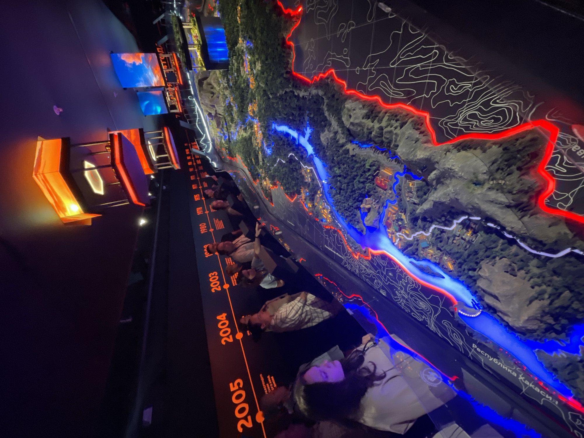 The image captures an exhibition hall, likely within the city of Krasnoyarsk, Russia, displaying a detailed diorama of a landscape featuring a river, mountains, and settlements. A timeline with years ranging from 2005 to 1952 is visible along the top left of the frame, illuminated in orange, indicating historical context for the diorama. Several people are observing the exhibit, their faces partially visible and illuminated by the ambient lighting and the exhibit itself. The diorama is enhanced with blue lights tracing the river and red lights outlining topographical features or boundaries. Above the diorama, large screens display colorful images, possibly depicting skies or conceptual representations, adding to the immersive experience of the exhibition. The overall environment is dark, emphasizing the illuminated display.