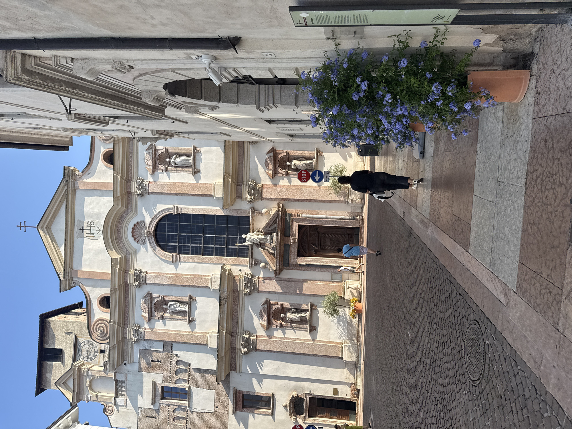 The image depicts the exterior of the Duomo of Trento, a Romanesque cathedral located in Trento, Italy, on a clear day. The foreground shows a cobblestone plaza with a circular metal grate. To the right of the plaza, a woman in black pants and a black top walks away from the camera, carrying a white bag over her shoulder. She is walking on a tiled pathway adjacent to a building. A large terracotta pot overflows with vibrant blue flowers, positioned on the right side of the pathway. Further down the pathway, a few other people are visible, walking in various directions. The Duomo itself, with its intricate facade and towering clock tower, dominates the background. The architecture features a blend of stone and light-colored stucco, adorned with statues and decorative elements. Sunlight casts shadows across the plaza and the building, indicating a bright day.
