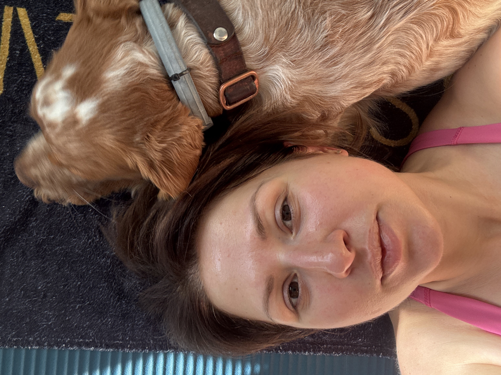 A person with fair skin, dark brown hair, and wearing a pink top is lying on a black mat. A reddish-brown and white dog with a brown leather and gray metal collar is lying partially on the person's head. The edge of a light blue surface is visible at the bottom of the image. The location is not identifiable as Sofia, Bulgaria, based on the provided image.