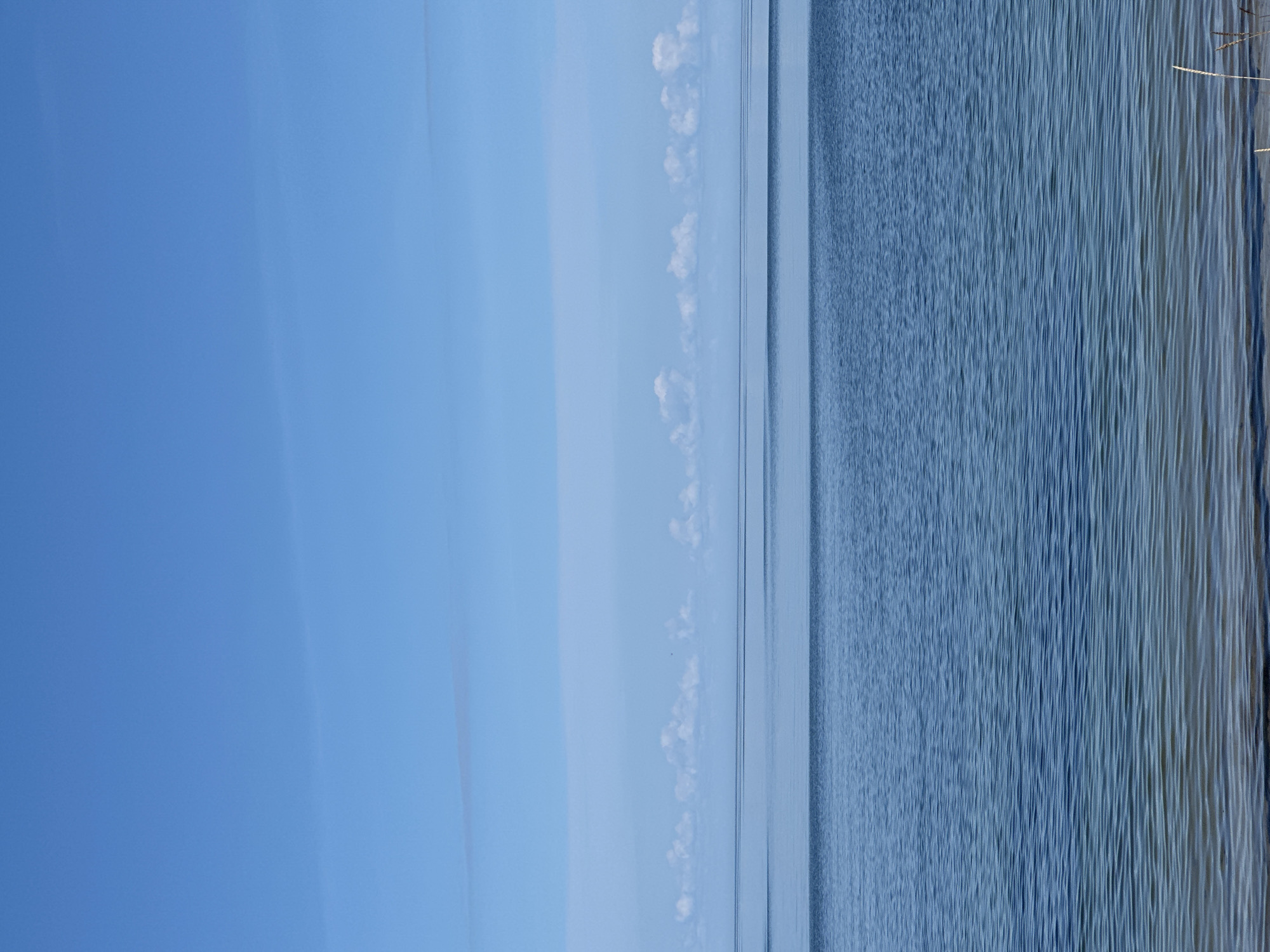 The image depicts a serene natural landscape at Meremõisa, Estonia. The scene is dominated by a vast expanse of calm blue water, likely a bay or sea, meeting a clear blue sky. The horizon line is sharp, with a thin band of lighter blue clouds stretching horizontally across the sky. Closer to the water's surface, a row of small, fluffy white clouds is visible. The foreground shows the edge of the water with subtle ripples and a sandy or pebbly shore, with a few dry reeds or grasses protruding. The overall impression is one of tranquility and open space. There are no people or man-made objects prominently featured in the image.