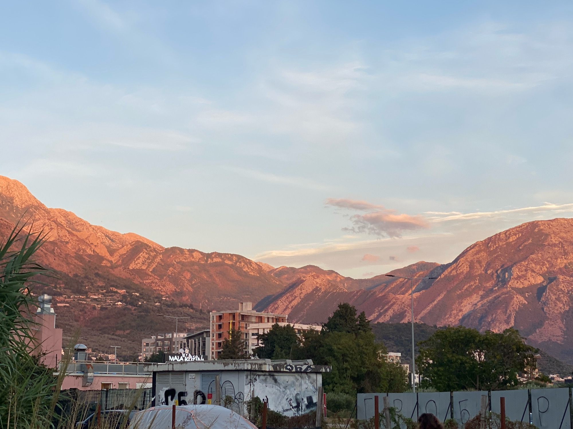 In Bar, Montenegro, a scenic landscape unfolds with rugged mountains bathed in the warm, golden light of sunset. The mountains dominate the background, their slopes dotted with scattered buildings, suggesting a town nestled within the natural beauty. In the foreground, a collection of urban structures, including buildings and what appear to be industrial or storage units, are visible. Some of these structures are adorned with graffiti, adding a layer of urban grit to the scene. Vegetation, including tall grass and the fronds of a palm-like plant, partially obscures the view of the foreground elements. The sky above is a soft gradient of blue and pink, with wispy clouds adding texture. A street lamp stands prominently in the middle ground, a testament to the presence of infrastructure within this transitional zone between nature and urban life.