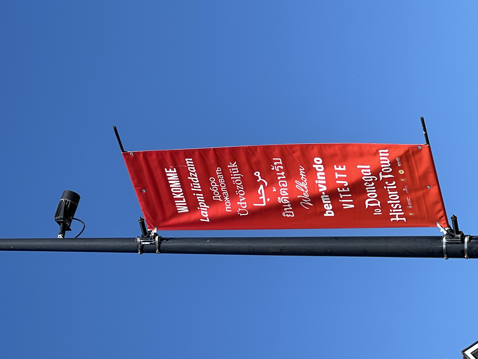 The image shows a bright red banner hanging vertically from a black lamppost against a clear blue sky. The banner displays the word "WILKOMME" at the top in large white sans-serif letters, followed by several lines of text in different languages, all in white. The languages include what appears to be Irish, Russian, Hungarian, Arabic, Thai, and Dutch. The bottom of the banner reads "to Donegal Historic Town" in a stylized white font. Below this, there are small logos, one of which is labeled "BALLAST". A black security camera is mounted on the lamppost above the banner. The setting is outdoors, and the lamppost is likely situated in a public area within Donegal Town, Ireland, given the text on the banner.