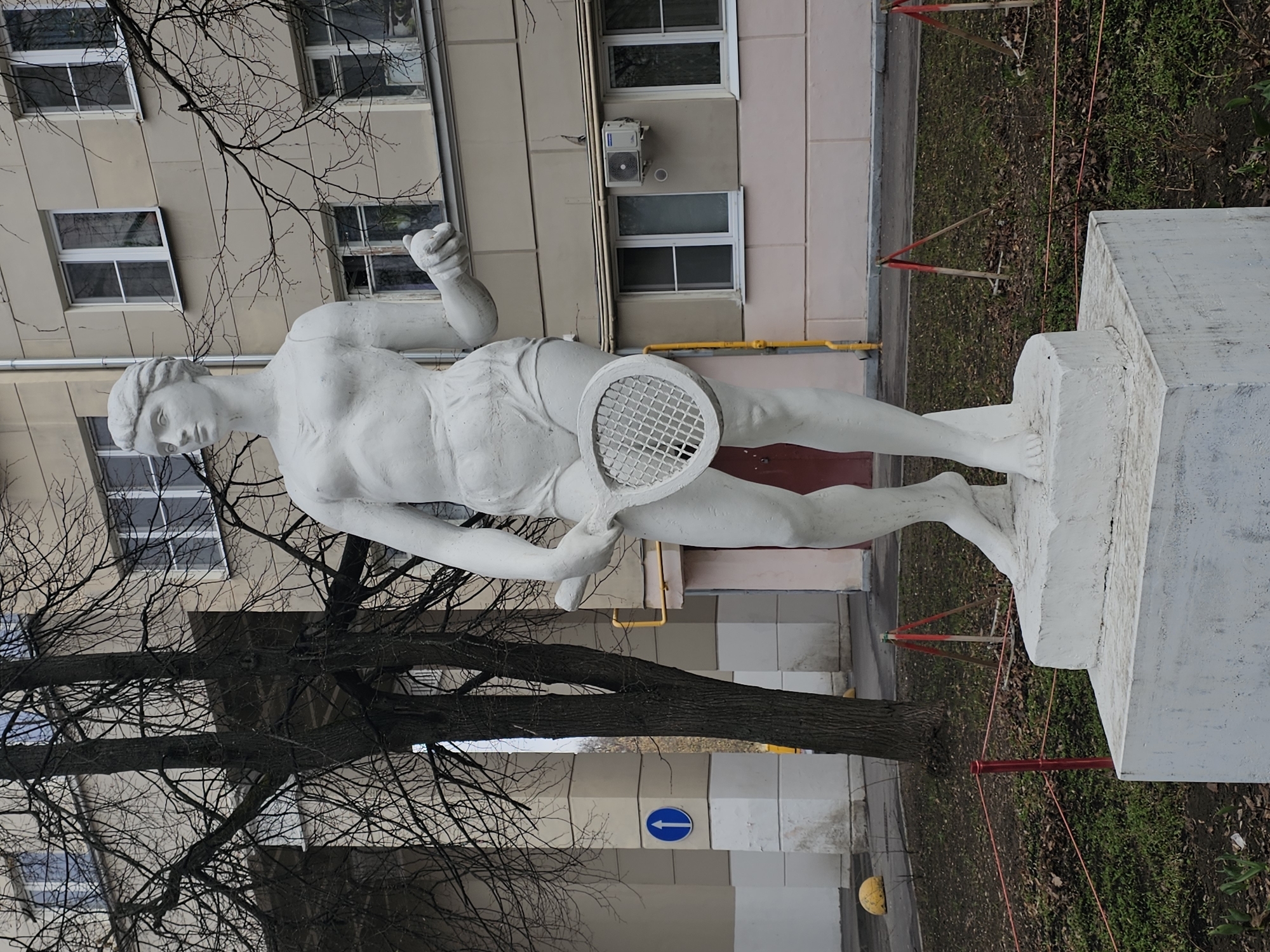 The image depicts a white statue of a tennis player in Khimki, Russia. The statue stands on a white pedestal amidst a backdrop of a building with multiple windows. A bare tree with thin branches partially obscures the view. The statue portrays a muscular figure with a tennis racket. The surrounding environment includes grass and fallen leaves. A yellow ball is visible on the ground near a building wall with a blue directional arrow. A red fence-like structure surrounds the statue.