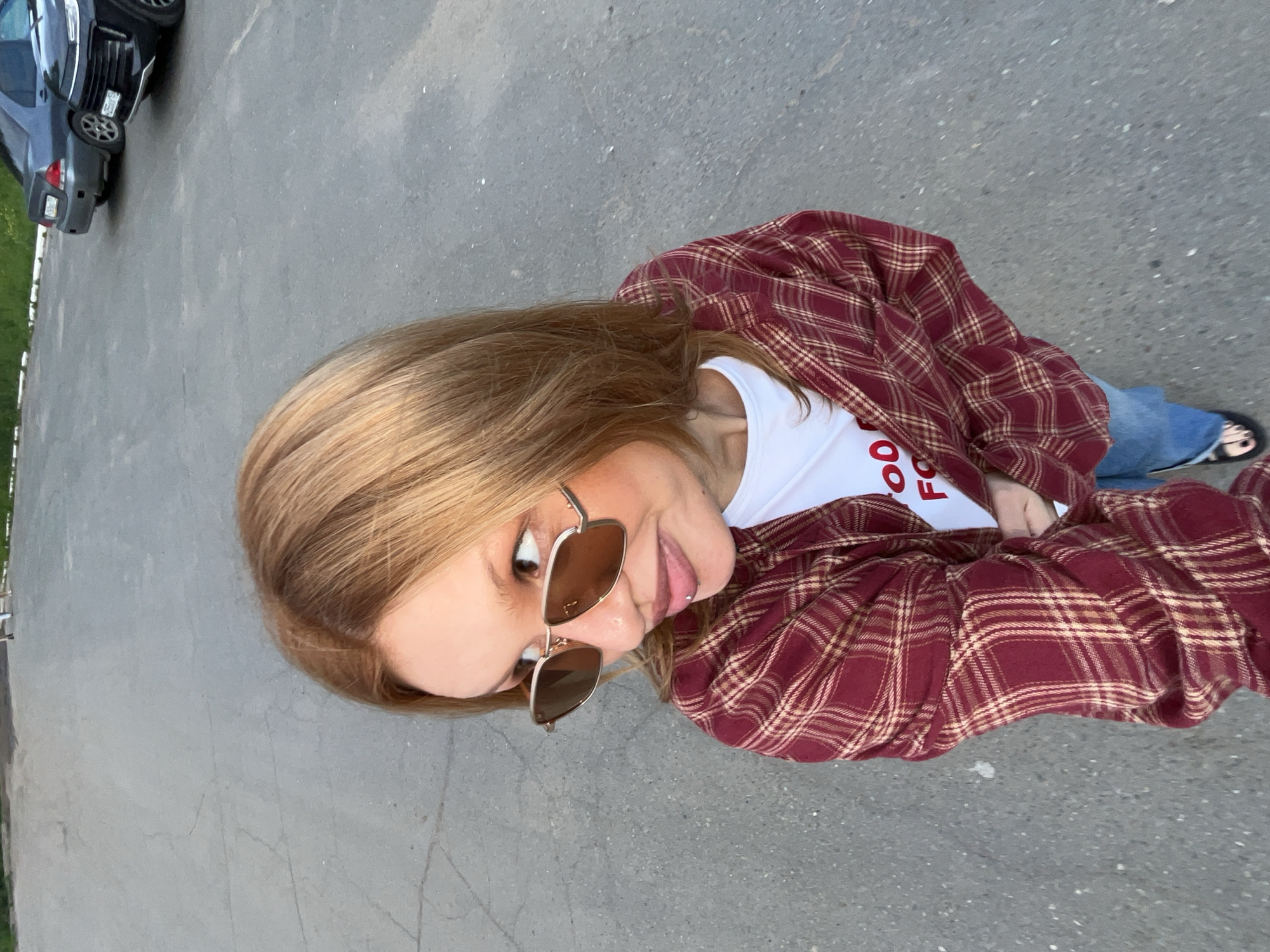 A young woman with auburn hair stands in a paved outdoor area, possibly a parking lot. She is wearing a red and tan plaid open shirt over a white t-shirt with red lettering, blue jeans, and black sandals. She is also wearing large, square-shaped sunglasses with brown lenses. Her head is turned to her left, and she appears to be looking off-camera. In the background, several cars are parked. The ground is asphalt with visible cracks and some small white pebbles. The lighting suggests it is daytime, with the sun casting some shadows. The scene is likely in the city of Chaikovsky, Russia, given the context.