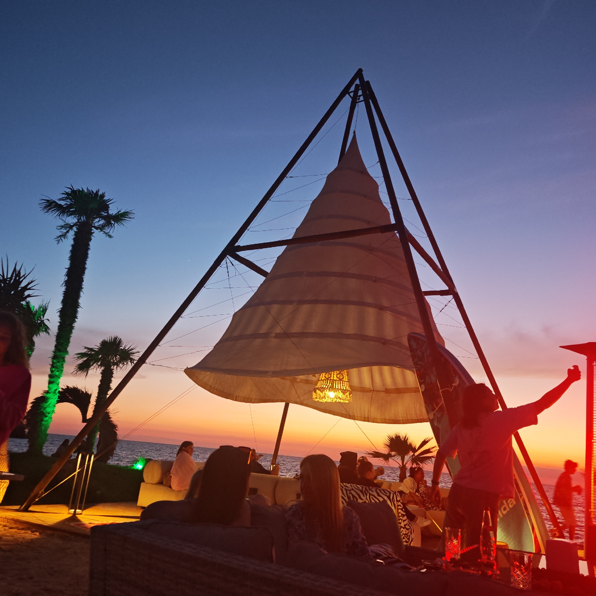 The image depicts a beachside scene at dusk in Мысовское сельское поселение, Ukraine. A large, sail-like structure with a woven lampshade hangs prominently in the center, supported by a metal frame. The sky transitions from deep blue at the top to orange and pink hues near the horizon, indicating sunset. Several palm trees are visible, some illuminated with green light.
In the foreground, people are seated on lounge furniture, facing the sea. Their backs are mostly to the camera. One person on the right, silhouetted against the sky, has their arm raised. To the left, a woman in a pink top is partially visible. Further back, other individuals are seated and appear to be socializing.
The environment suggests a relaxed, open-air gathering or bar area. The sea is calm and visible behind the people. Surfboards are propped up against the central structure. In the distance, the coastline and possibly buildings are faintly visible. The overall atmosphere is warm and inviting, characteristic of a beach setting during twilight.